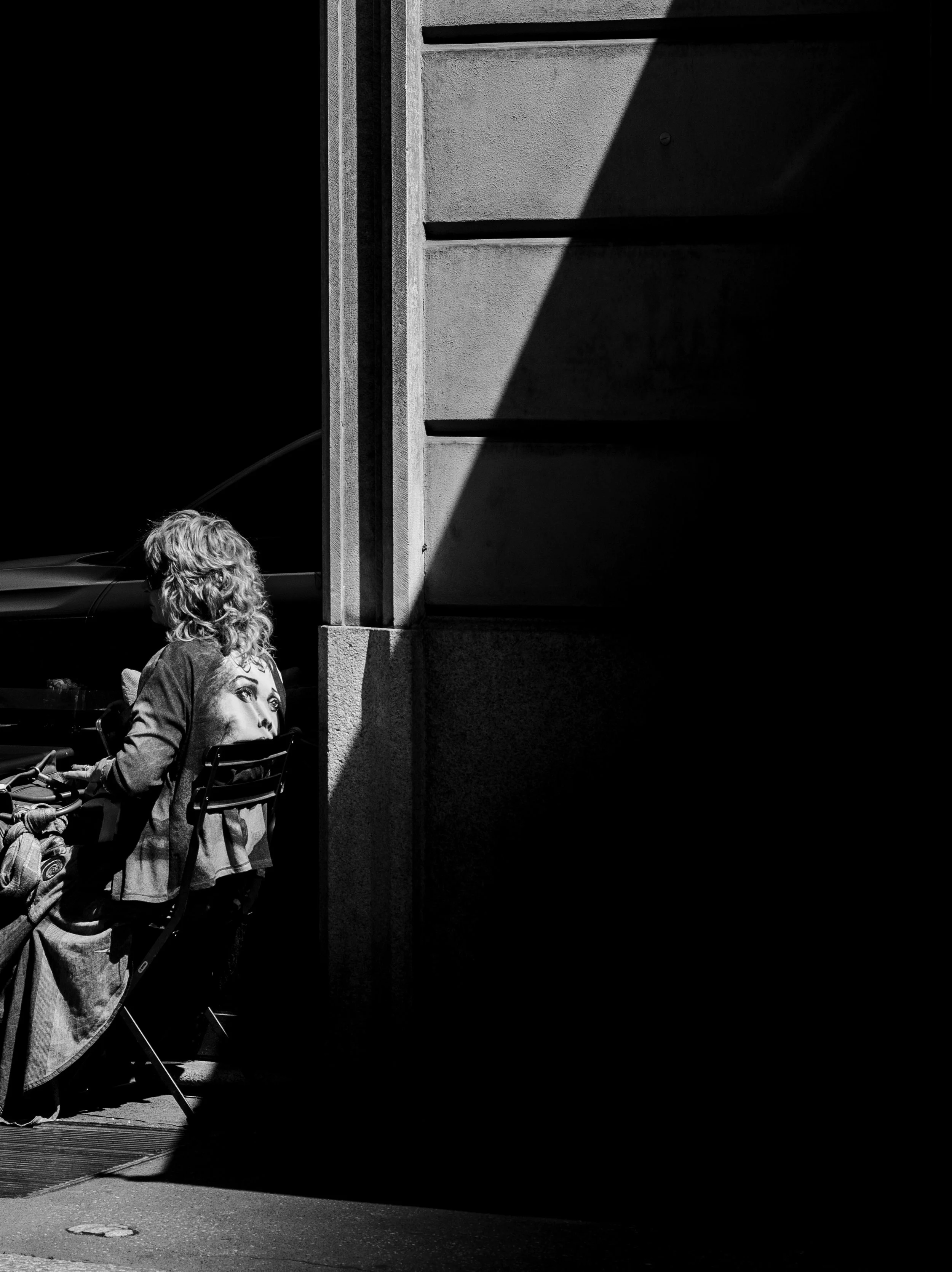 A black and white photo of a woman sitting on a bench with her back to the camera. She has curly hair and is wearing a jacket with a photo of a woman's face on the back. There is a tent and a sidewalk in the foreground and a building wall with shadow