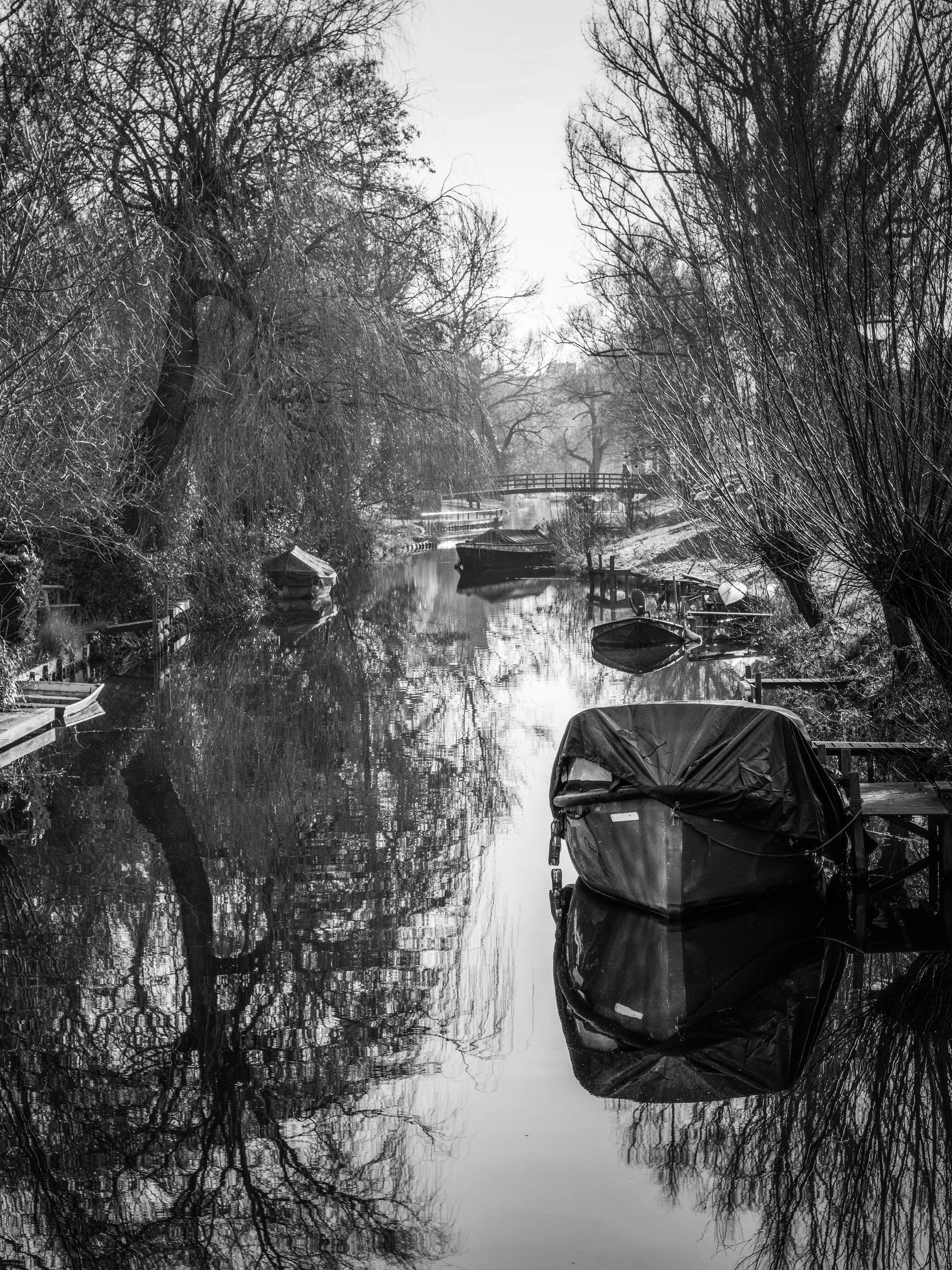 A black and white photo of a narrow river or canal with boats moored along the sides, surrounded by leafless trees, a bridge in the distance, and the reflection of the trees in the water.