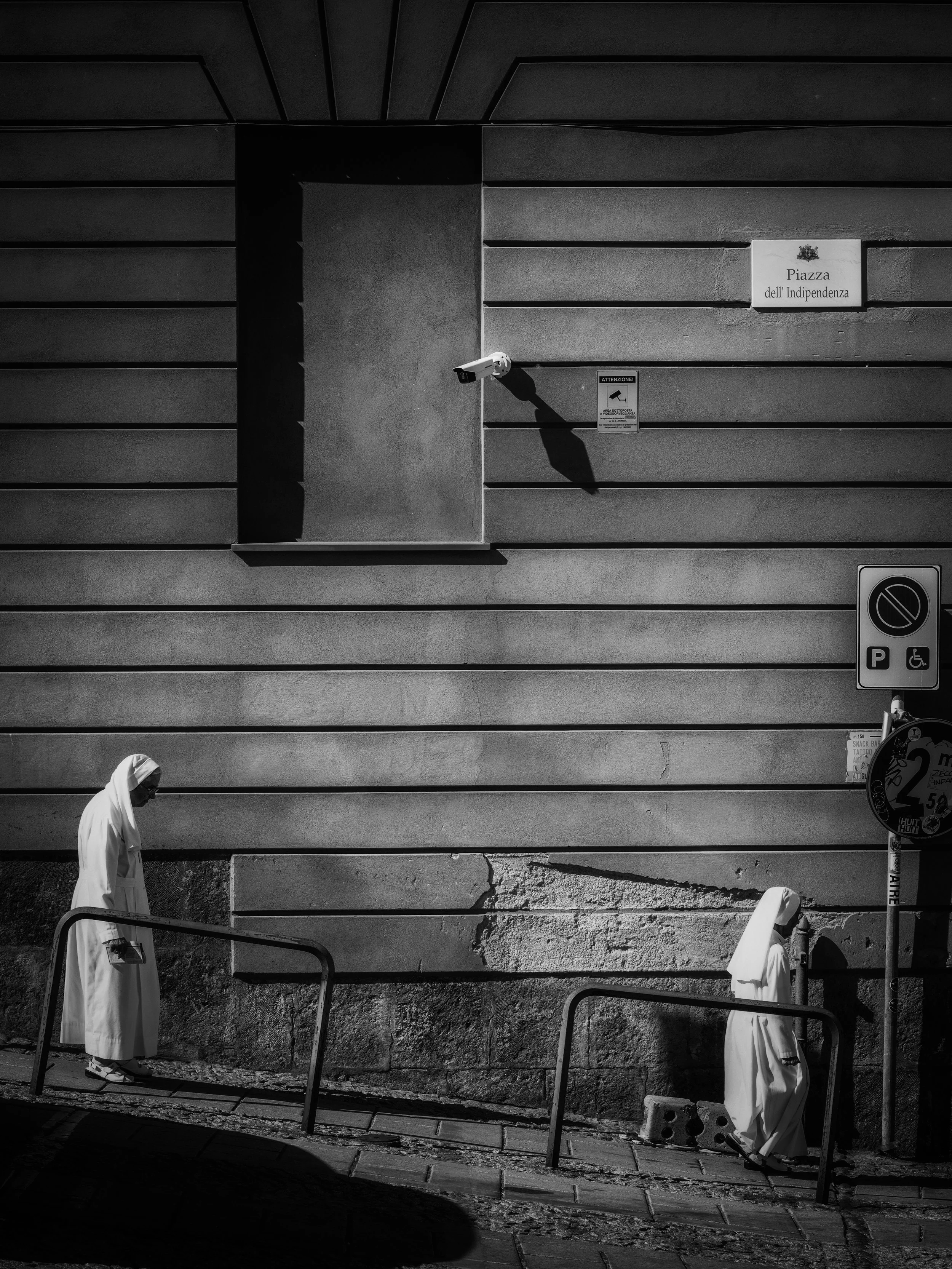 A black and white photo of a city street scene. Two women dressed in robes are walking past a building with horizontal siding. One woman is in the foreground, wearing sunglasses and white sandals, looking downward. The other woman is further back, mo