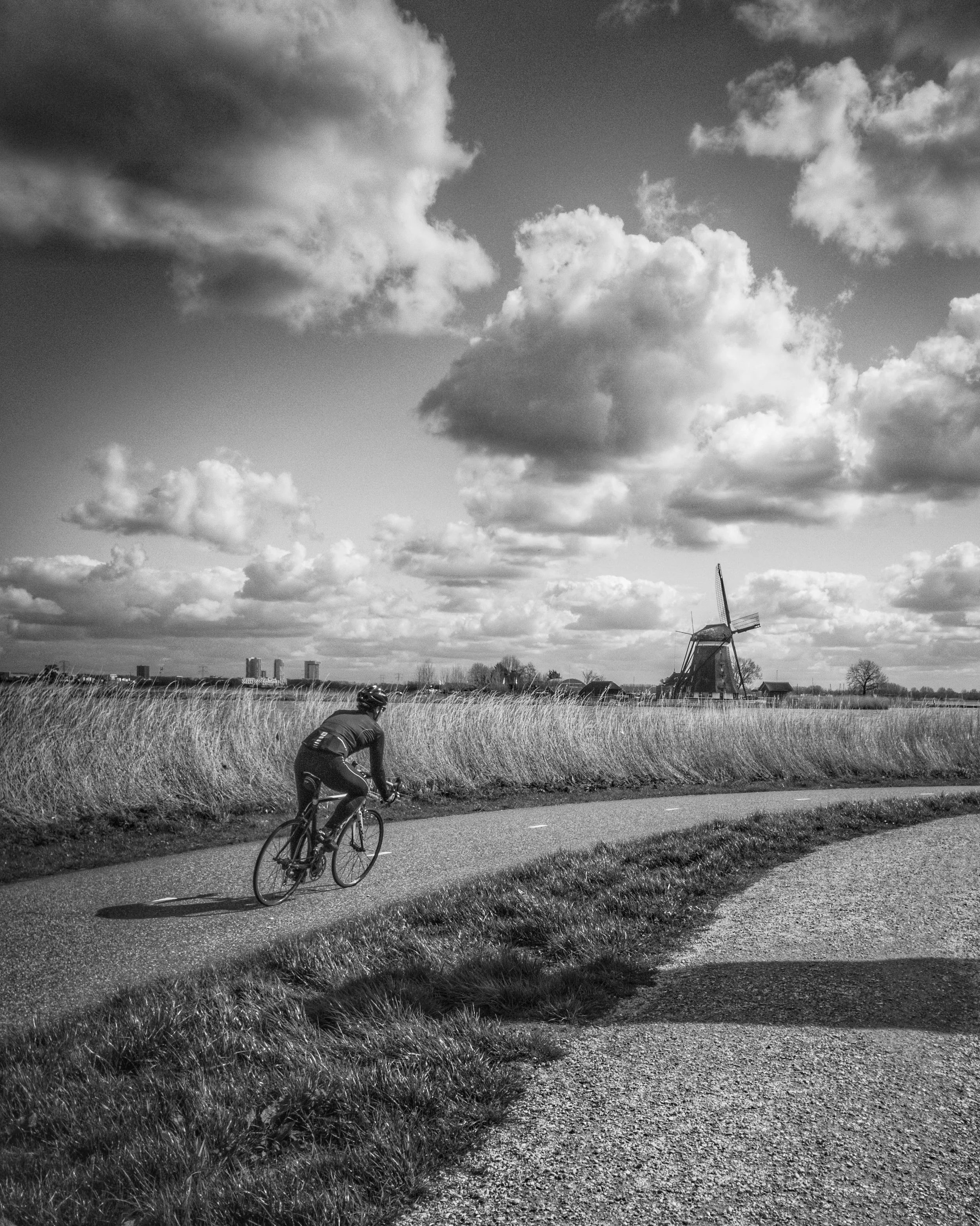 A black and white photograph of a rural landscape featuring a cyclist riding on a paved path, tall grass on the side, a traditional windmill in the background, and a sky filled with scattered clouds.