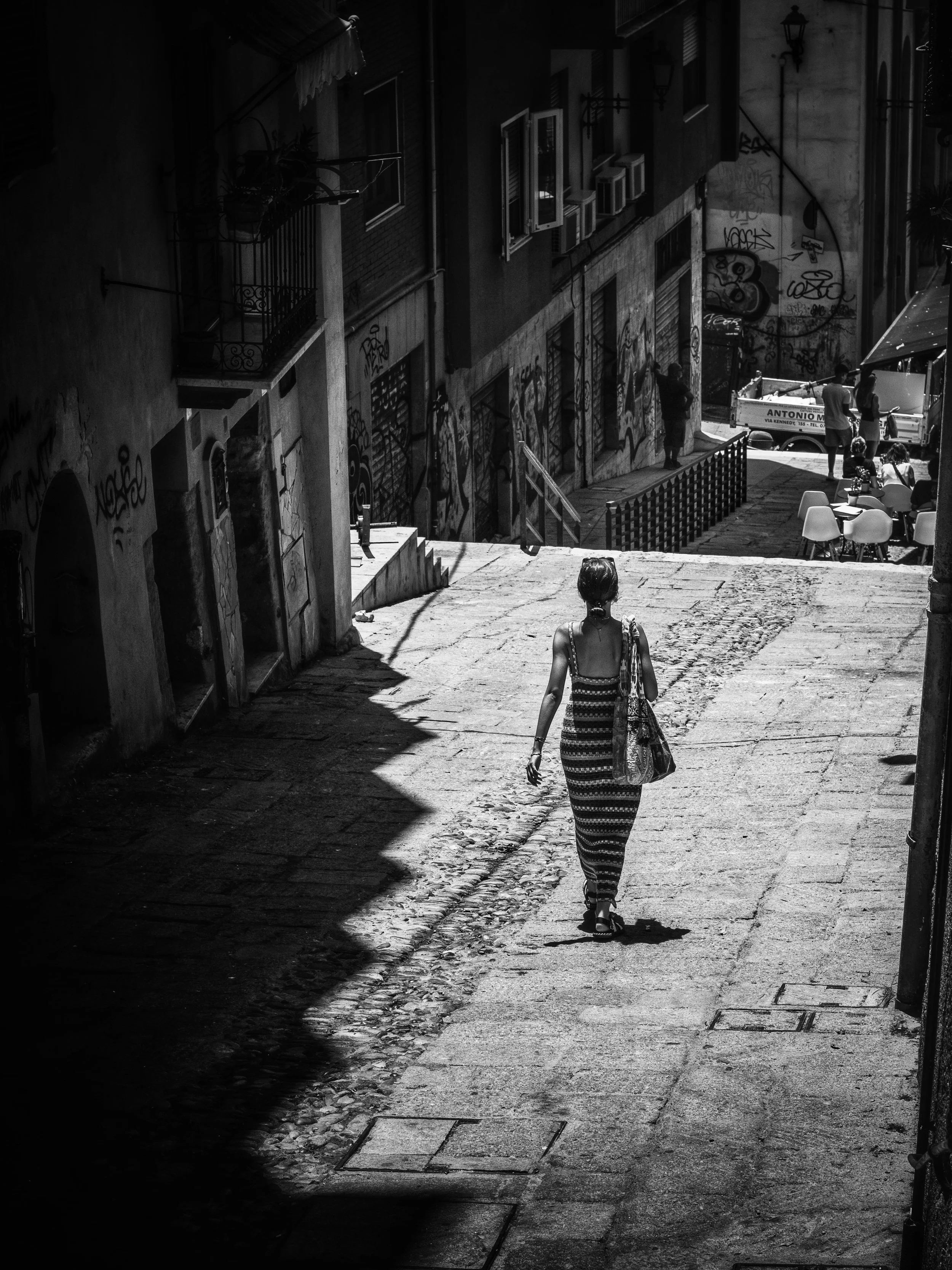 A woman in a striped dress walking down a narrow alleyway with graffiti-covered walls, sunlight creating shadows on the ground, and outdoor seating areas with people gathered.