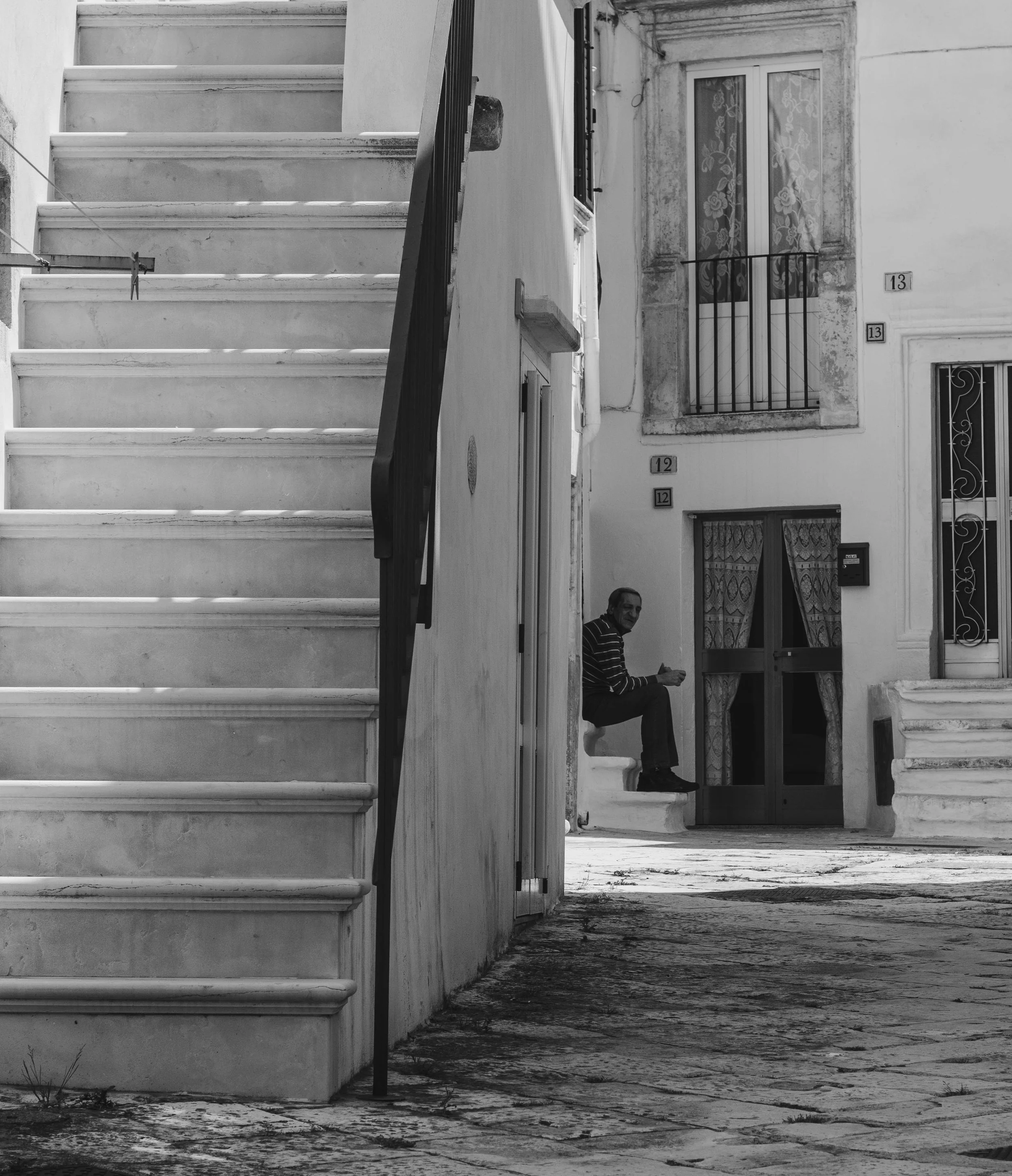 A black and white photo of a man sitting on a small stool on a sidewalk in front of a building, looking at something in his hand, with part of a staircase and a window with curtain in the foreground.