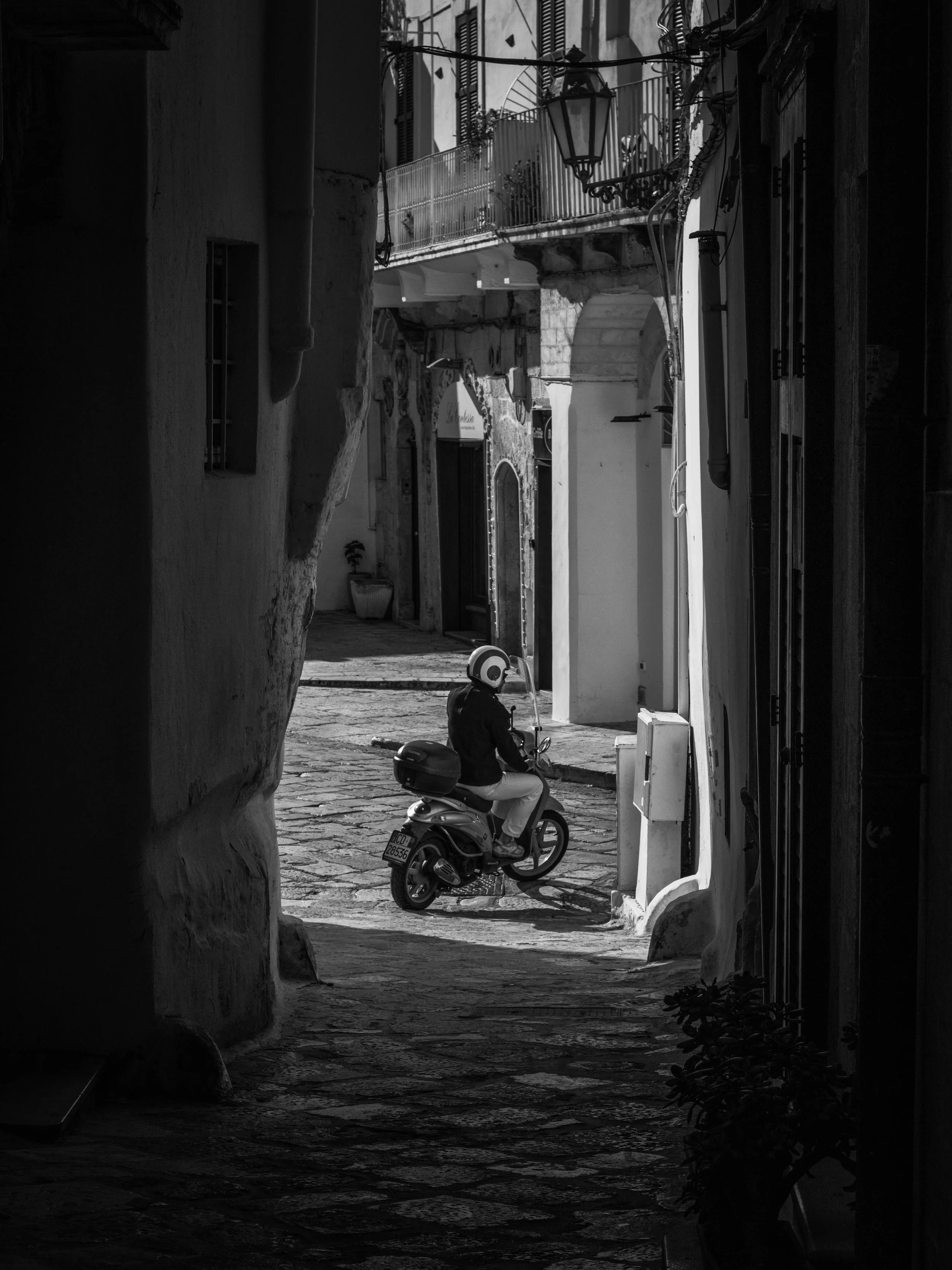 A narrow street scene in black and white, with a person riding a scooter through an arched alleyway. The street is paved with cobblestones, and there are buildings with balconies and windows along the sides.