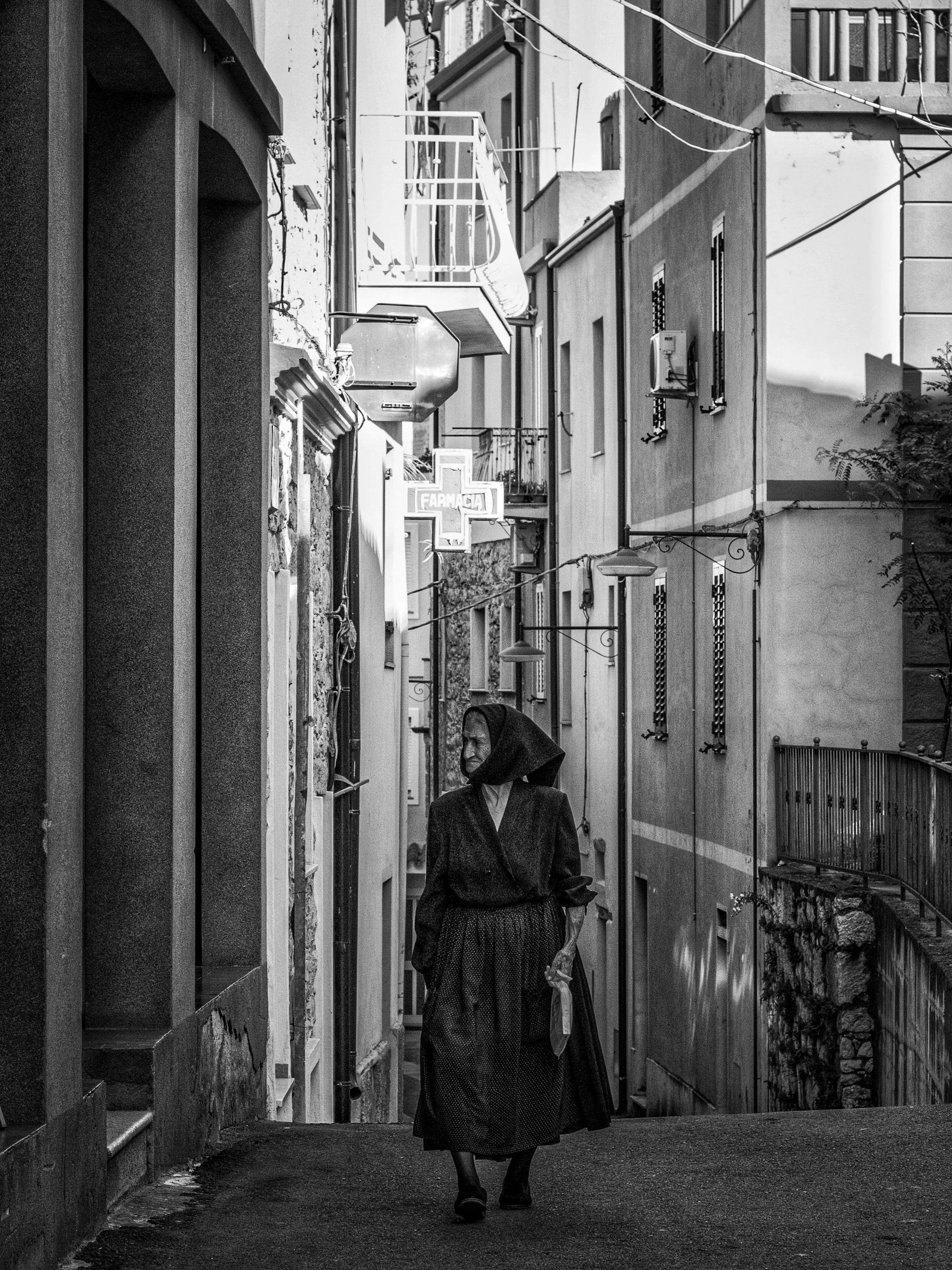 A woman walking along a narrow alleyway lined with buildings on either side, with a pharmacy sign visible in the background.