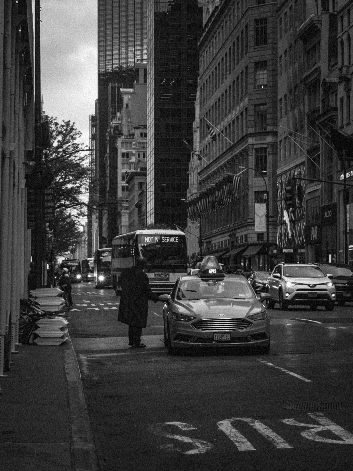 City street scene in black and white with tall buildings, cars, and a bus displaying "Not in Service." A person is walking on the sidewalk, and there are awnings and flags on the buildings.