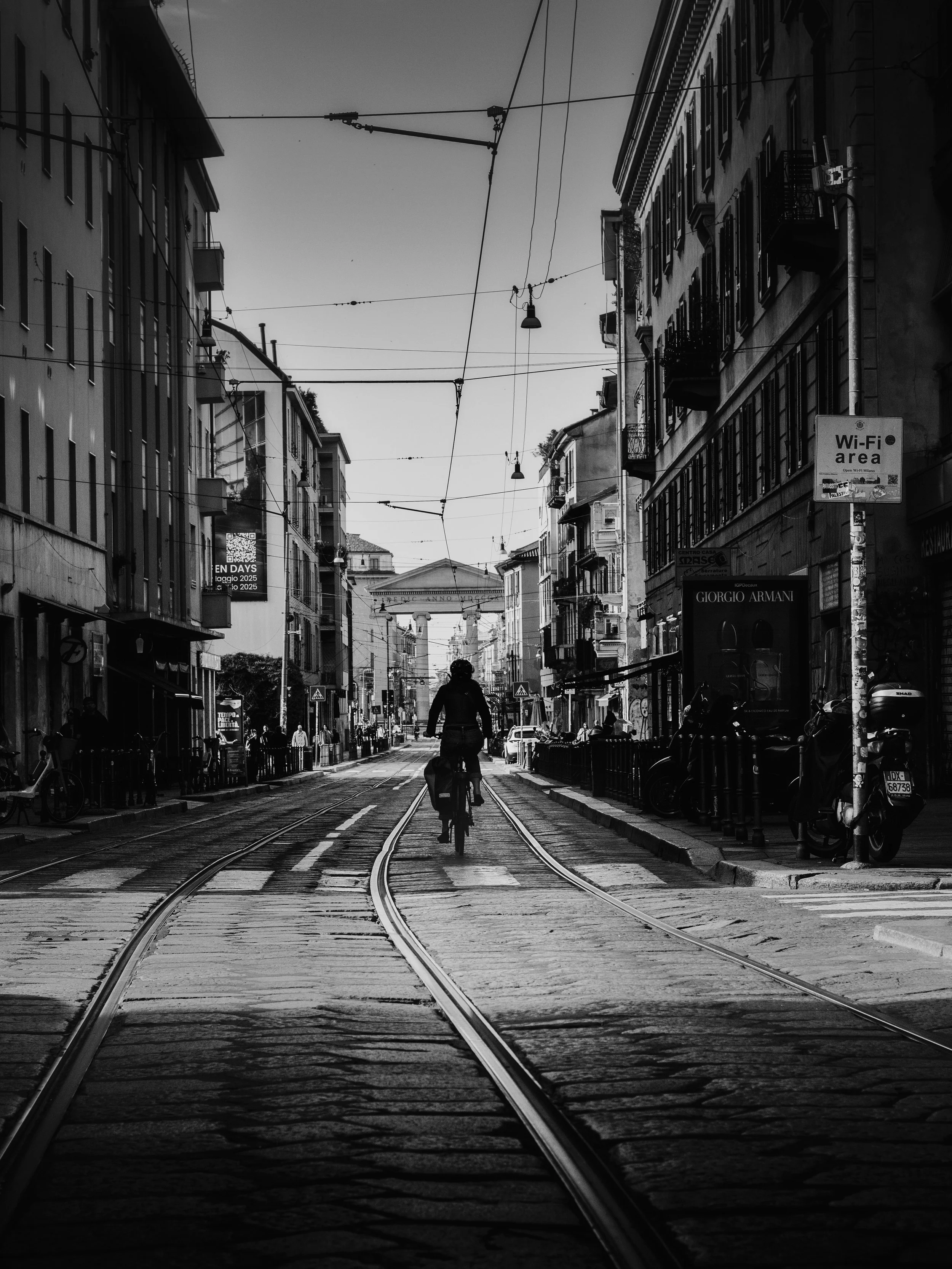 A black and white city street scene with tram tracks, buildings on both sides, a cyclist in the center, and overhead wires.