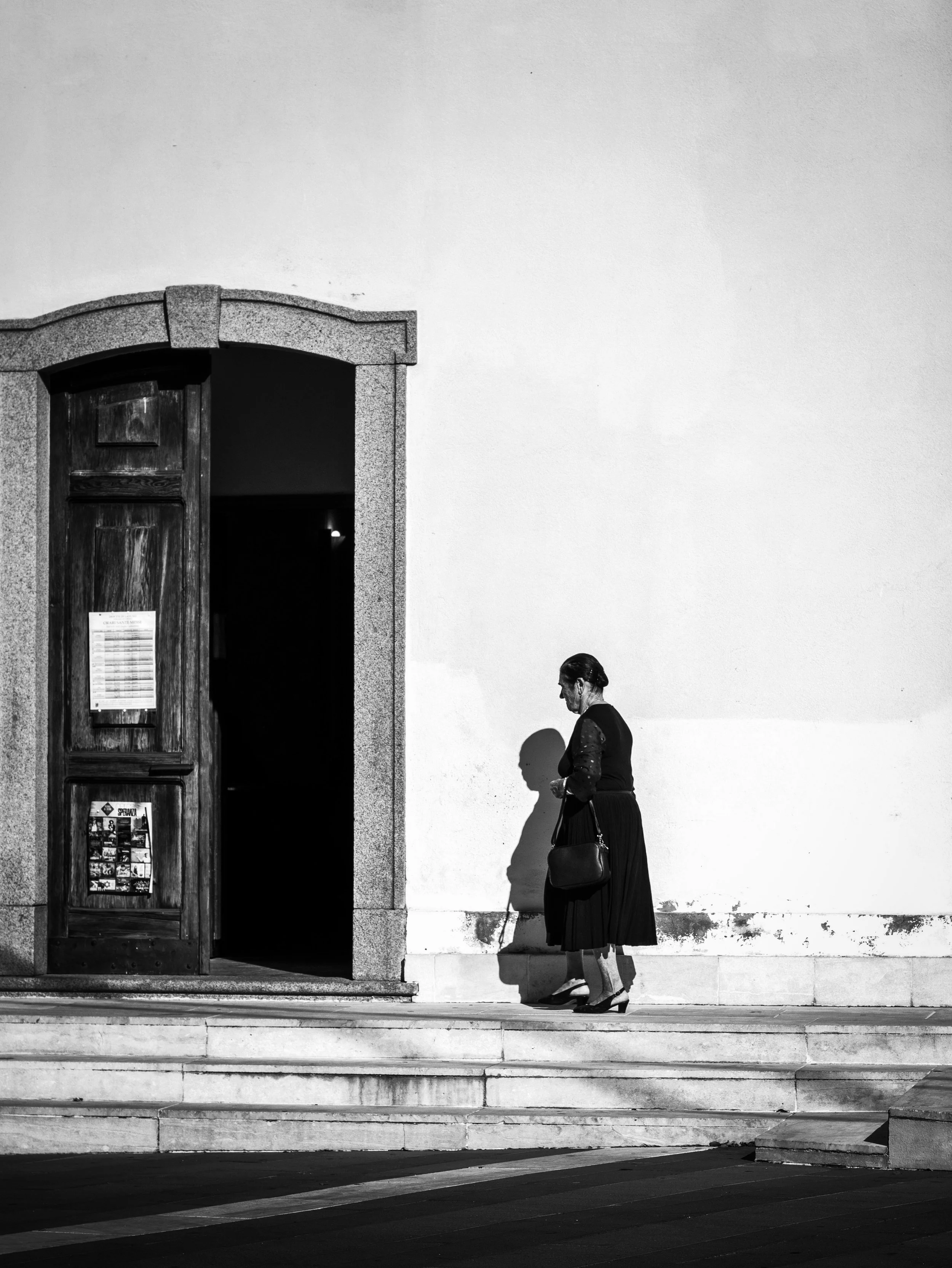 Black and white photo of a woman dressed in traditional attire standing against a plain wall near an arched door with posters. The woman is holding a purse and a cane, with her shadow cast on the wall beside her. There are three steps leading to the 
