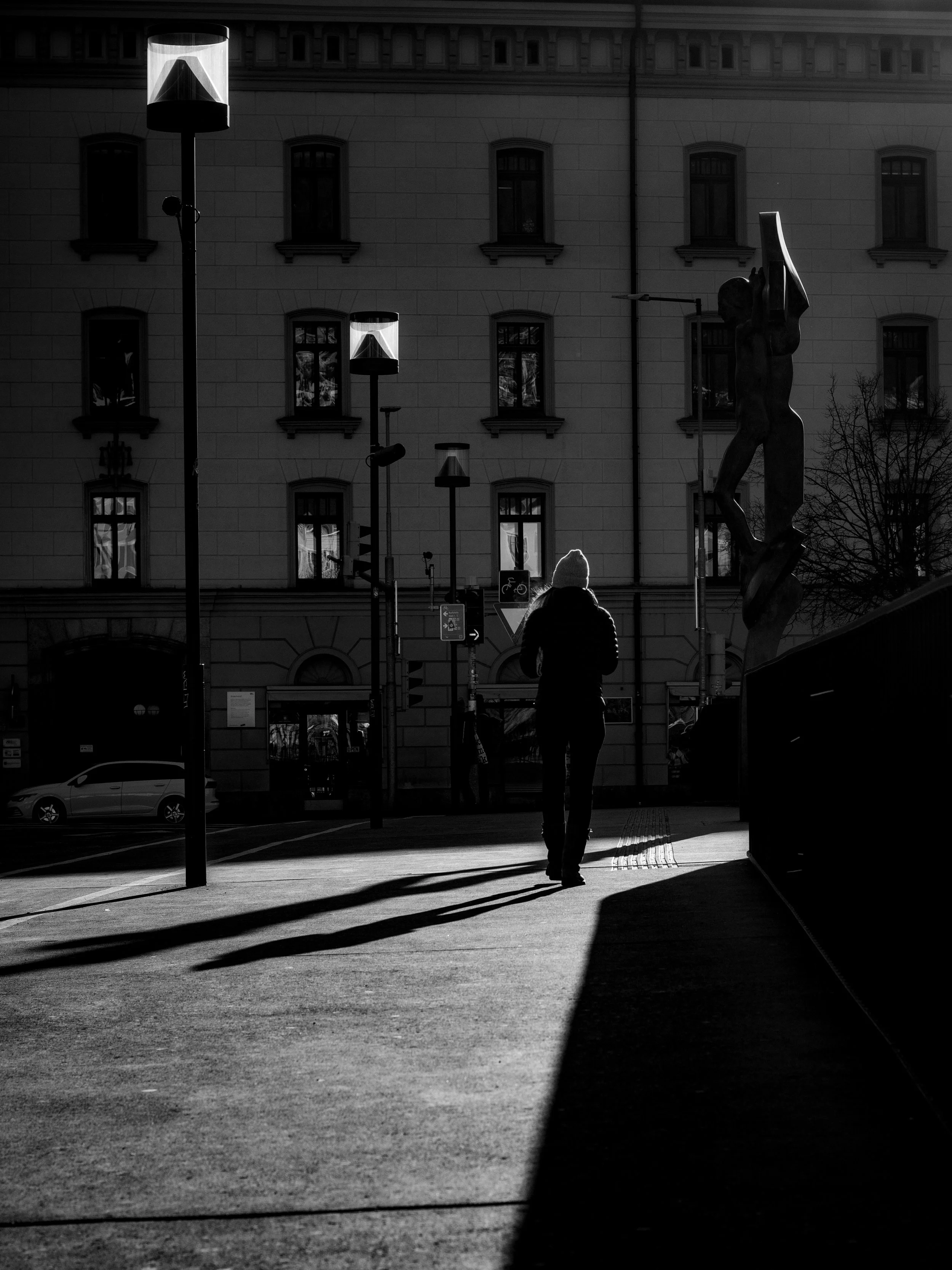 A person walking on a city sidewalk with a shadow cast by a large building and street lamps. The scene is in black and white, with a statue visible on the right and a building with multiple windows in the background.