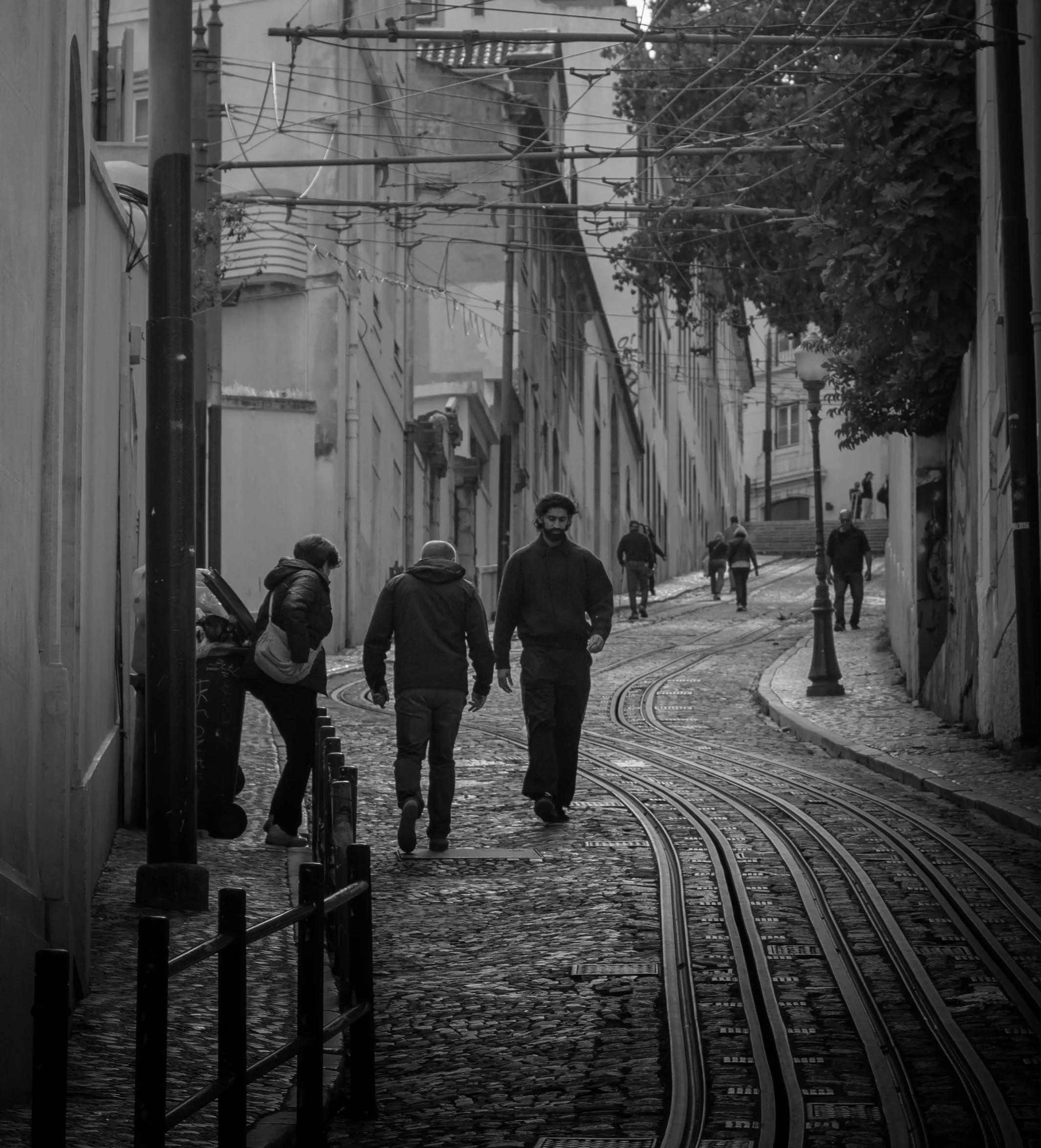 A black-and-white photo of a city street with uneven cobblestone pavement, tram tracks, and street lamps. Several people are walking on the street, some in conversation, with buildings and trees in the background.