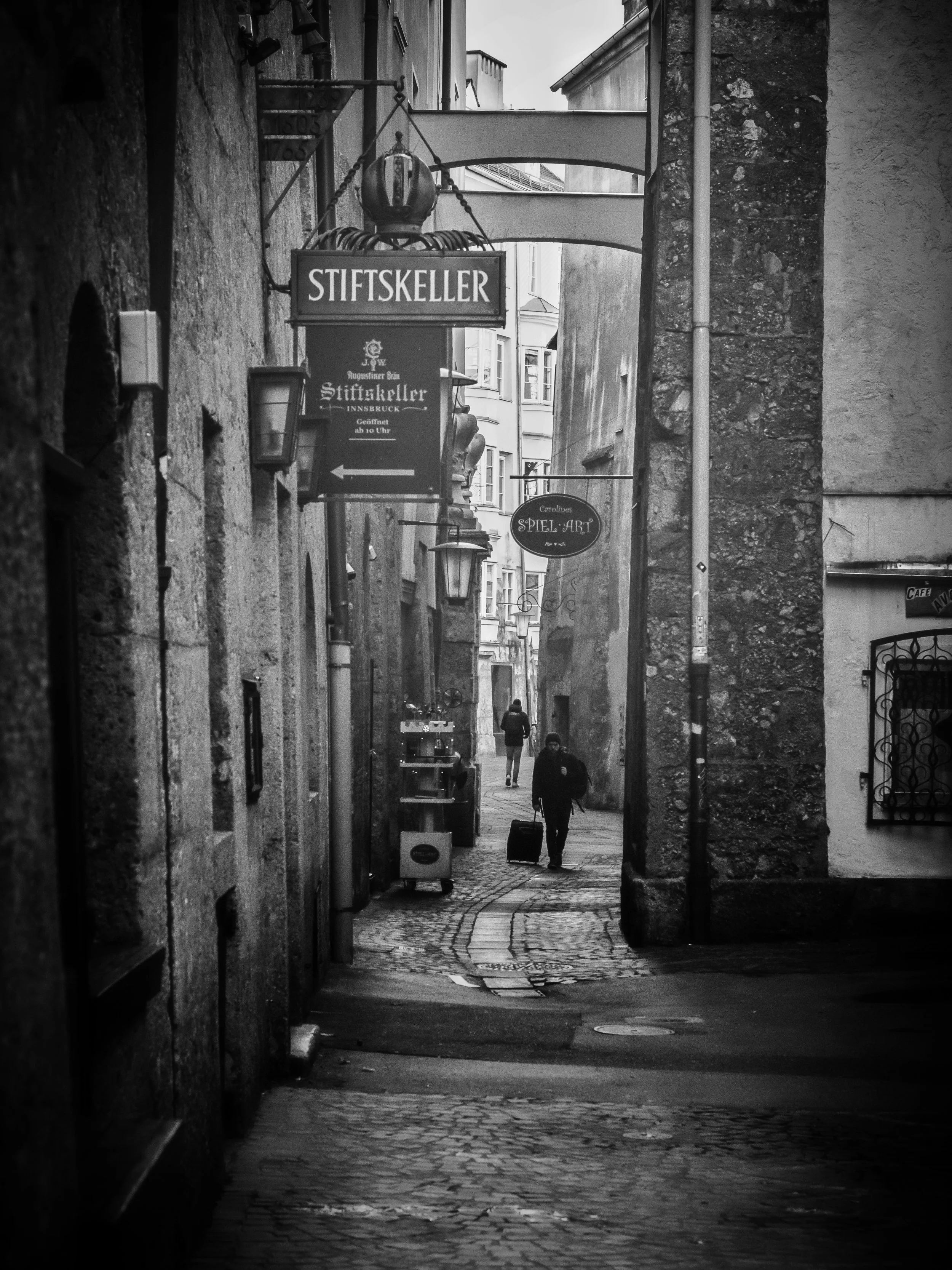 A narrow cobblestone street viewed through an archway, with a person walking with a suitcase in the distance, surrounded by old stone and plaster buildings, black and white photograph.