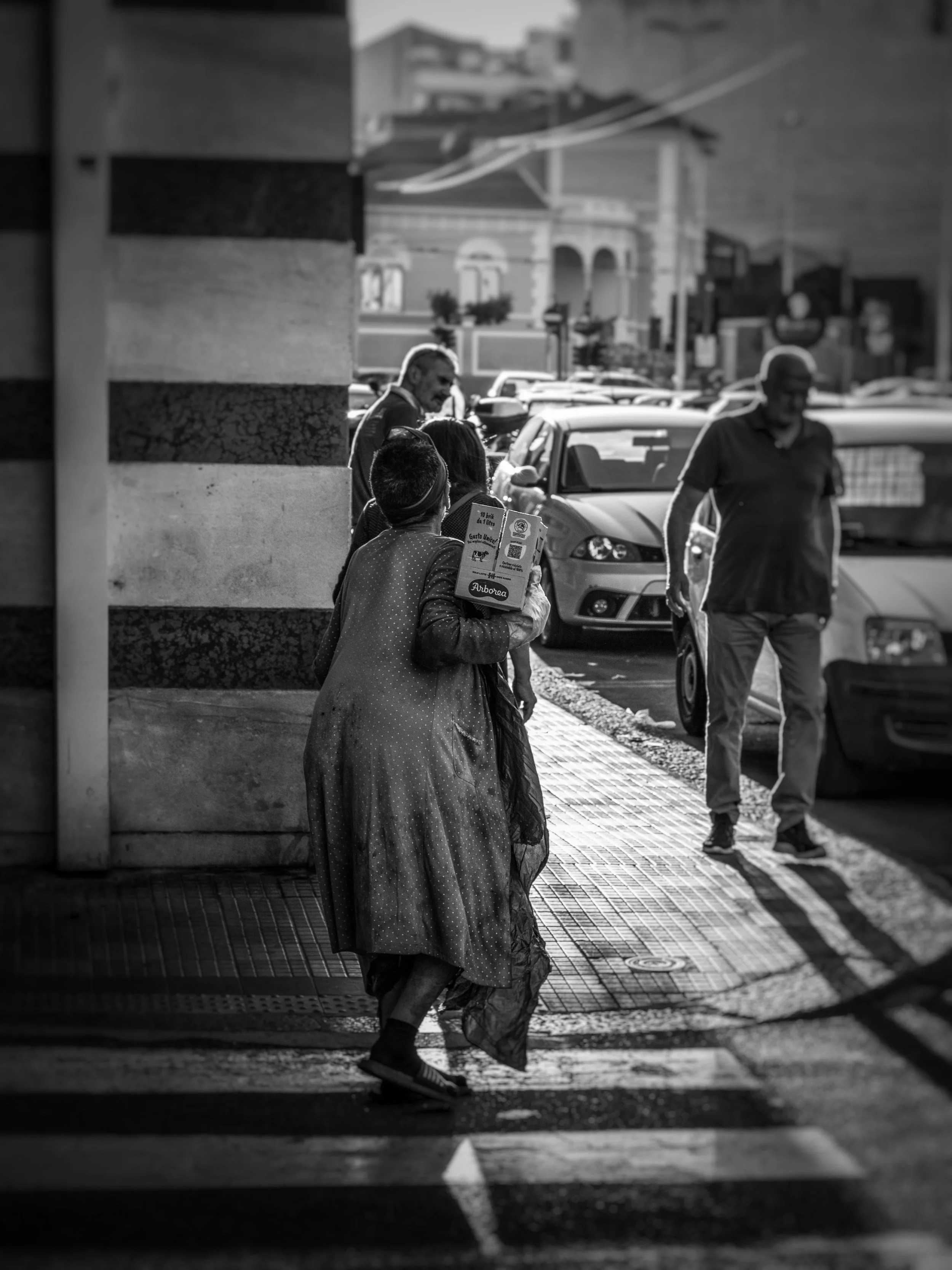 A black and white photo of a city street scene showing a woman in traditional attire and face mask walking on the sidewalk, with several cars parked and people walking, and a building with classical architecture in the background.