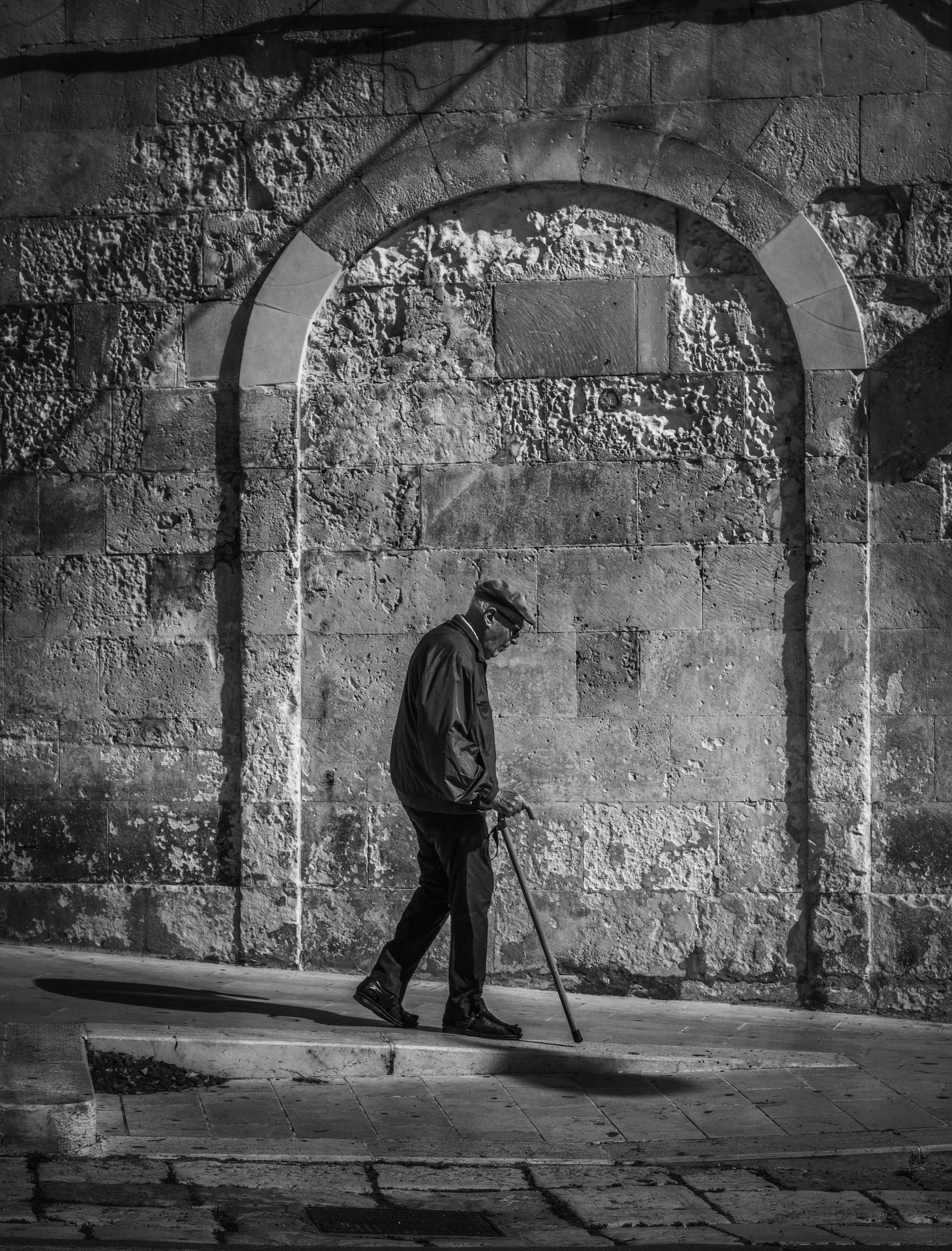 A black and white photo of an elderly man walking with a cane in front of a textured stone wall at night. Shadows from streetlights cast across the wall.
