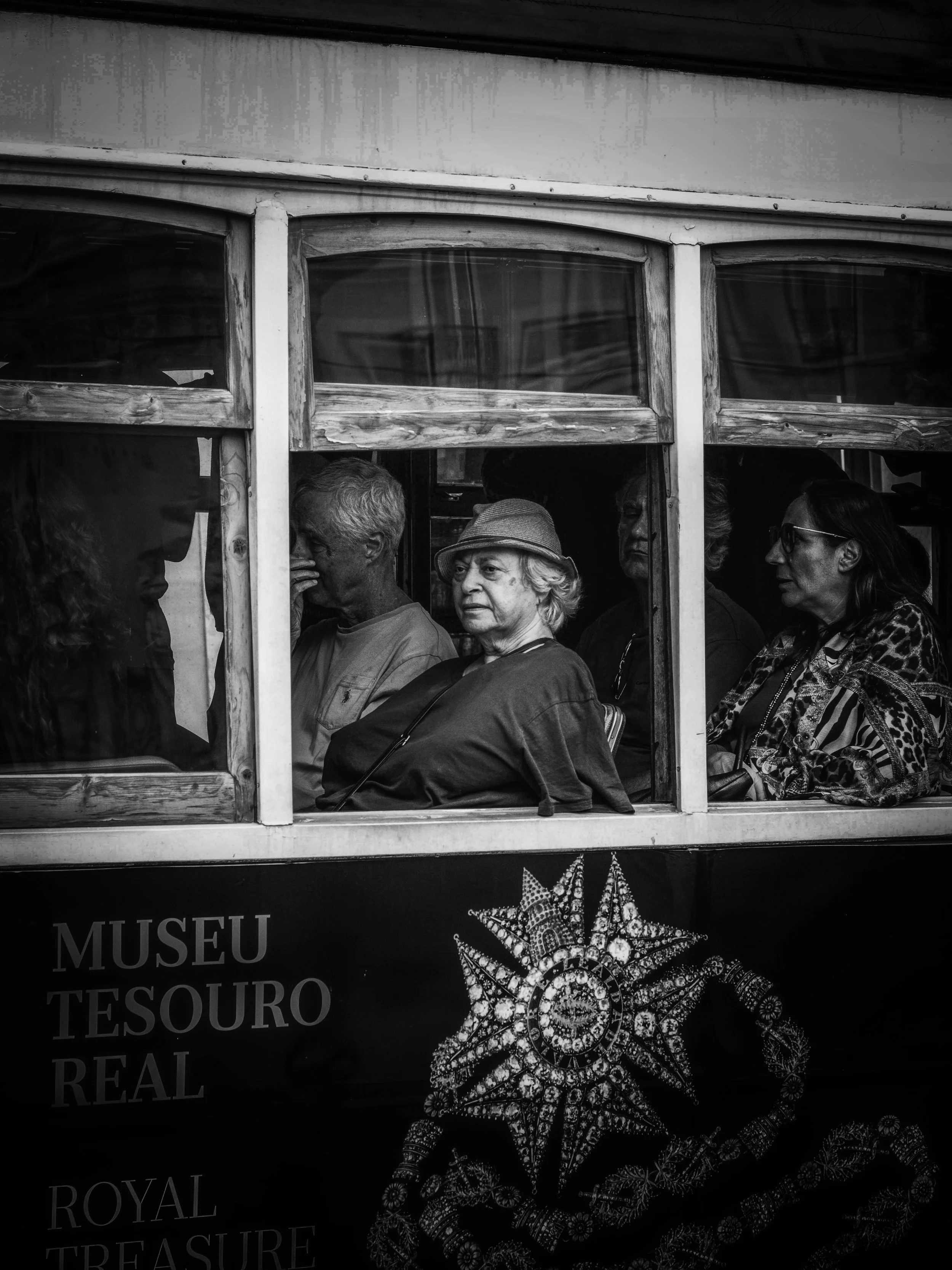Black and white photo of people sitting inside a bus viewed through the window. There are five women with different looks, one wearing glasses and another with a hat. The background features a bus interior and a sign below the window that reads "MUSE
