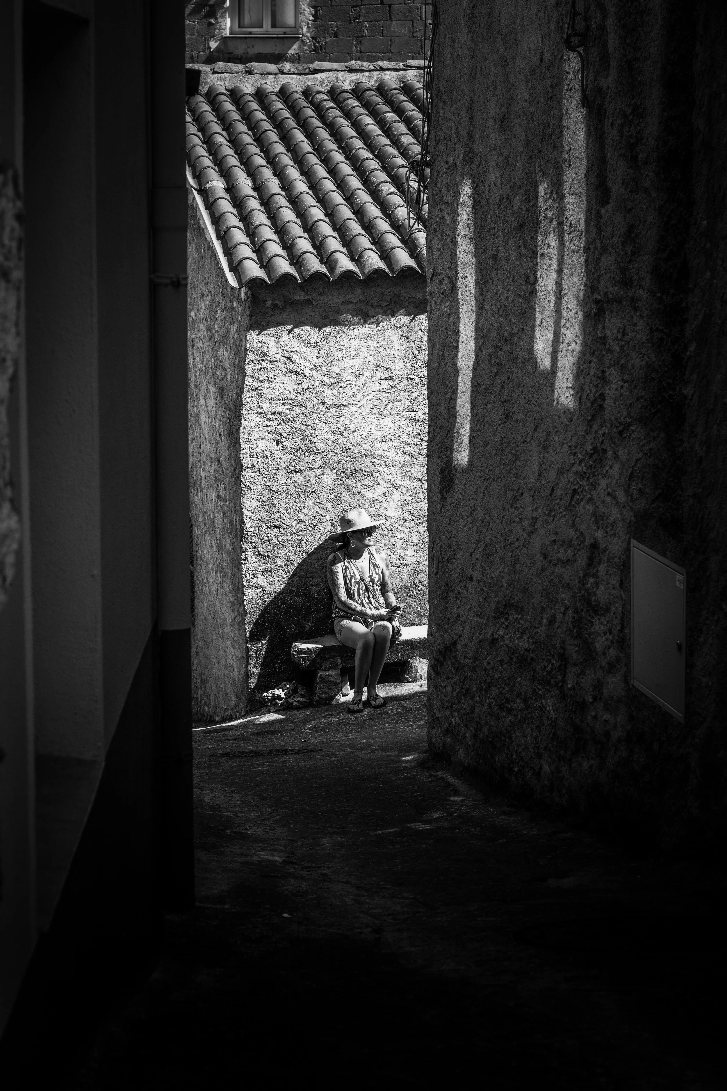 A woman sitting on a stone bench in a narrow alleyway with textured concrete walls, sunlight casting shadows, and tiled rooftops with a small window in the background.