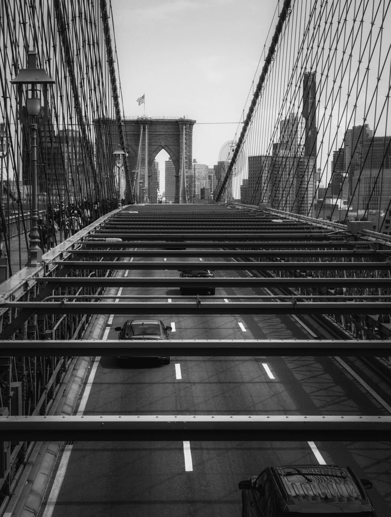 Black and white photo of the Brooklyn Bridge in New York City, showing the bridge's towers and cables, with a view of the city skyline and a road with parked cars below.