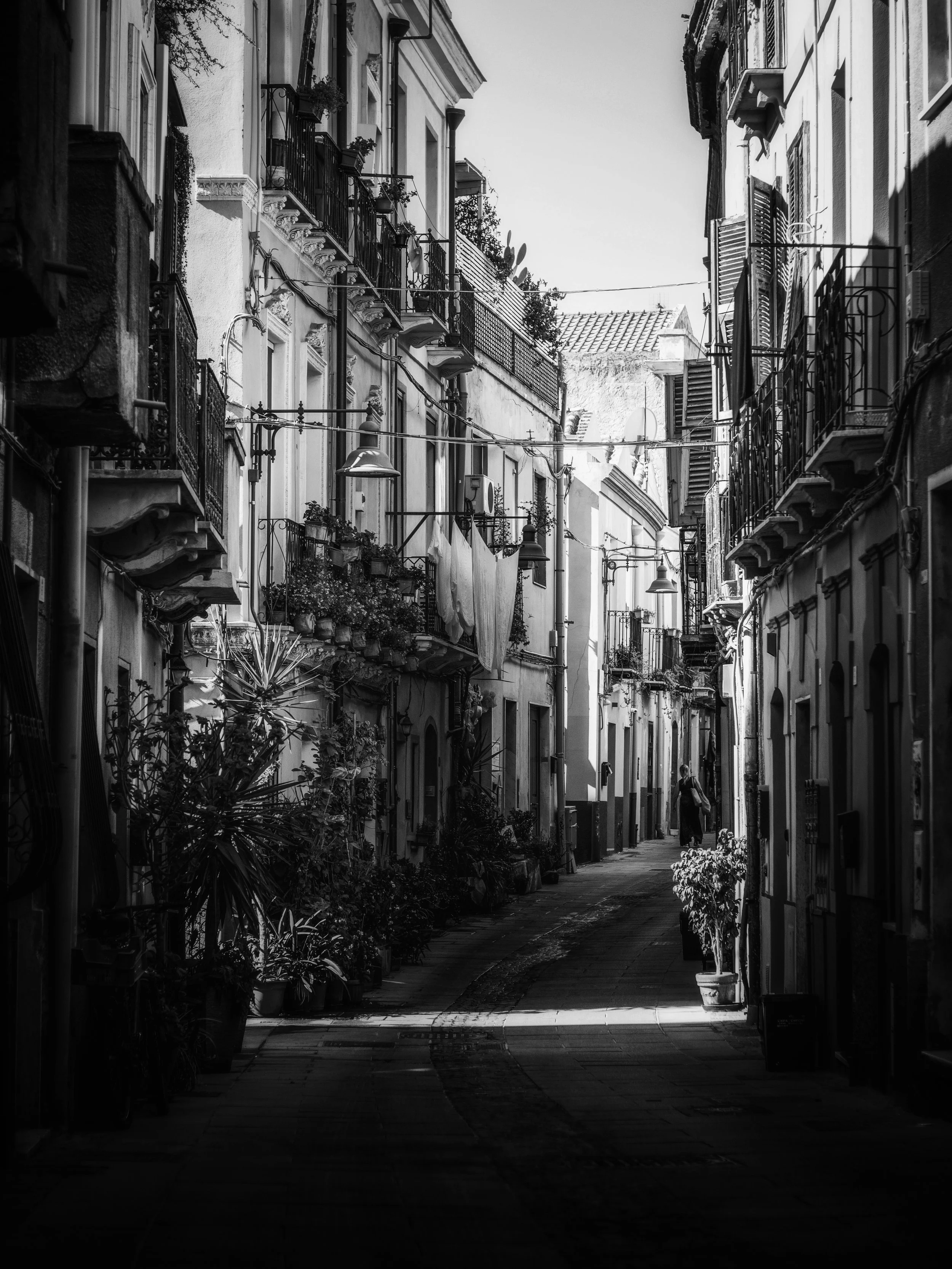 Black and white photograph of a narrow European-style street with balconies, potted plants, street lamps, and a person walking in the distance.