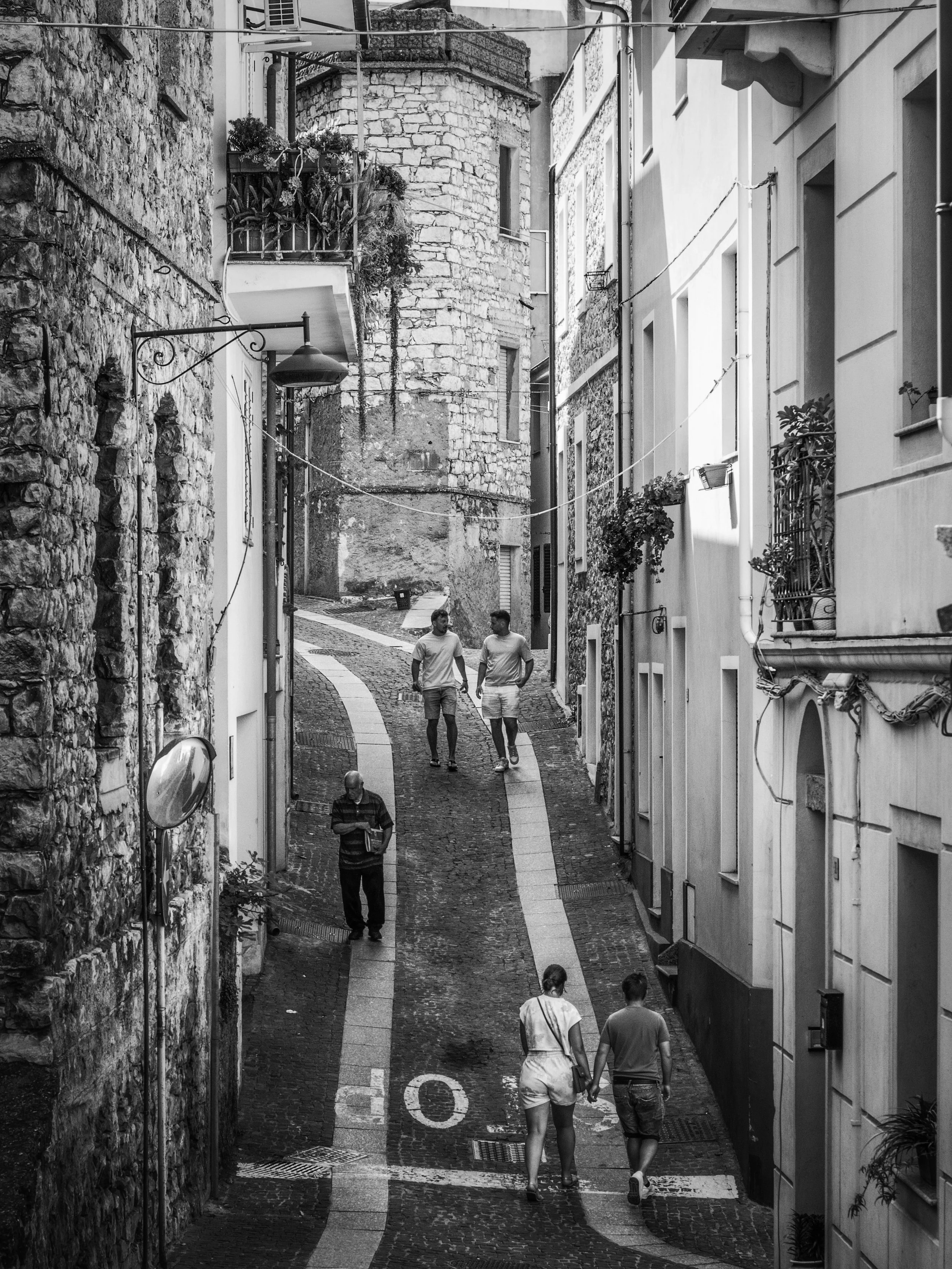 A grayscale image of a narrow, cobblestone city street with stone and plaster buildings. Several people are walking along the street, and some are holding hands. The street has signs, a streetlamp, and balconies with plants.