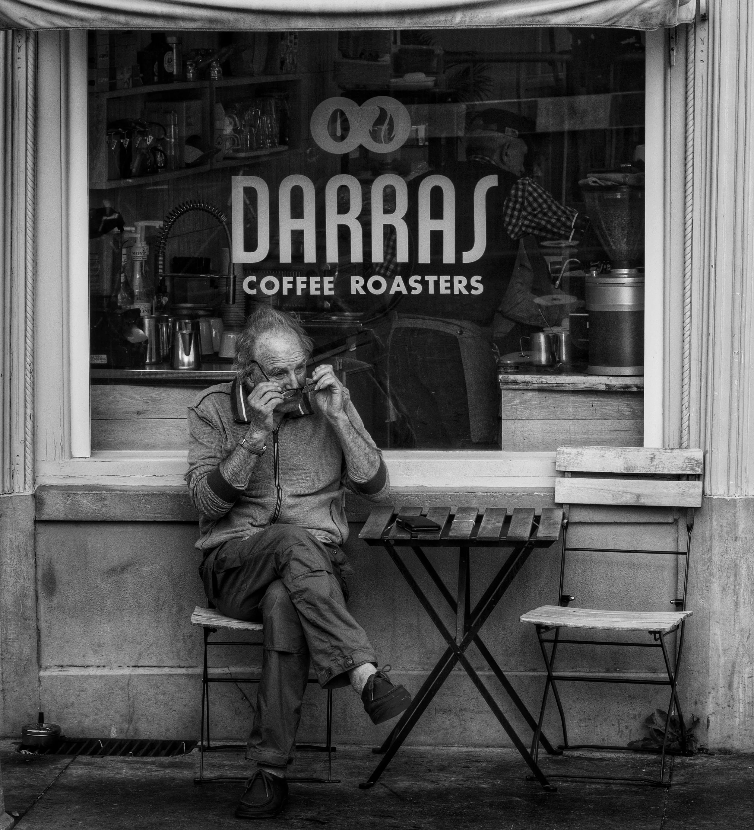 A man sitting on a wooden bench outside a coffee shop. The shop's window displays the logo '100 DARRAS COFFEE ROASTERS'. The man, wearing glasses, a jacket, and cargo pants, is adjusting his glasses and appears to be holding a face mask. The shop's i