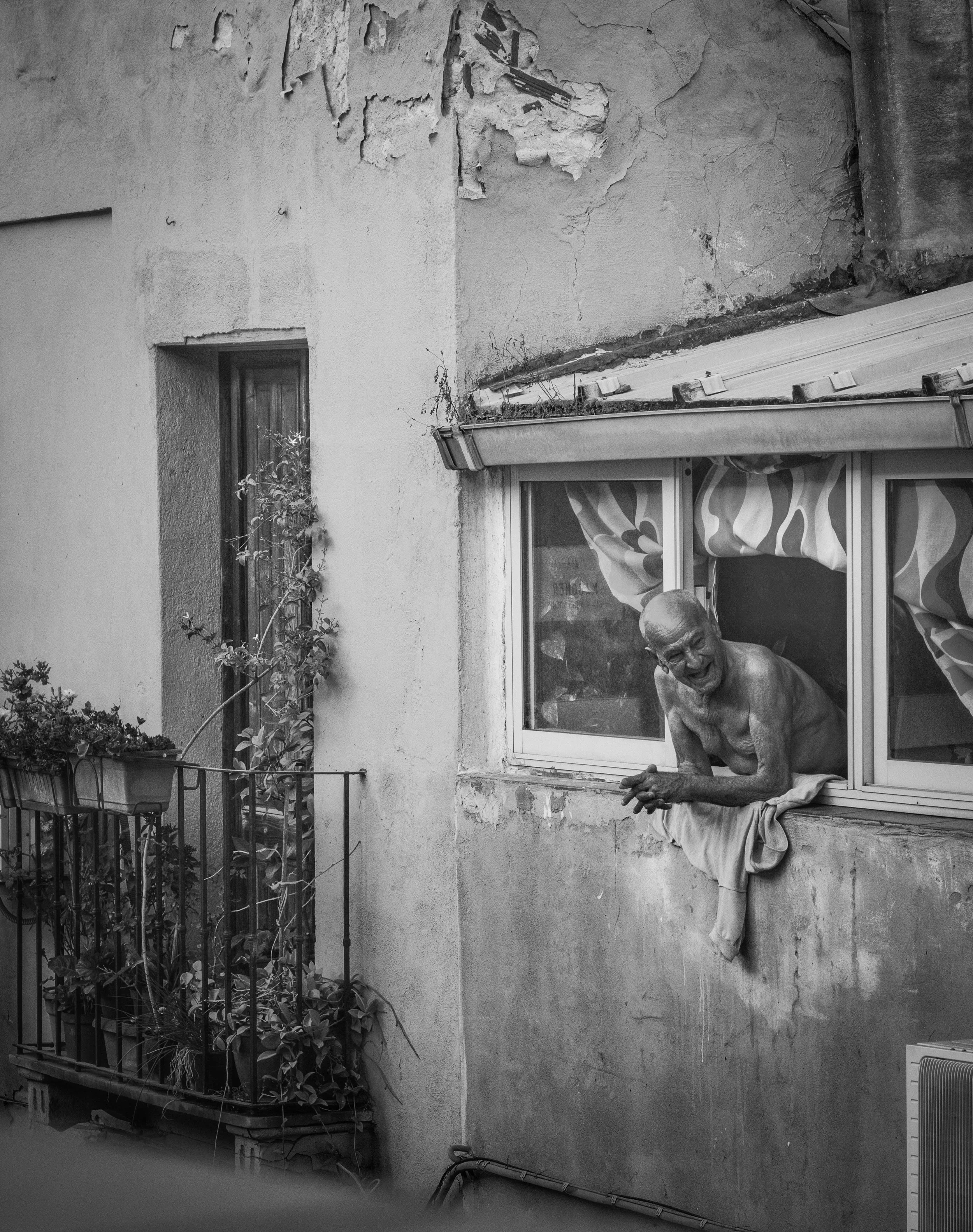 A shirtless elderly man leaning out of a window on a weathered building exterior, with peeling paint and exposed cracks, next to a balcony with plants and barriers, and an air conditioning unit on the ground.