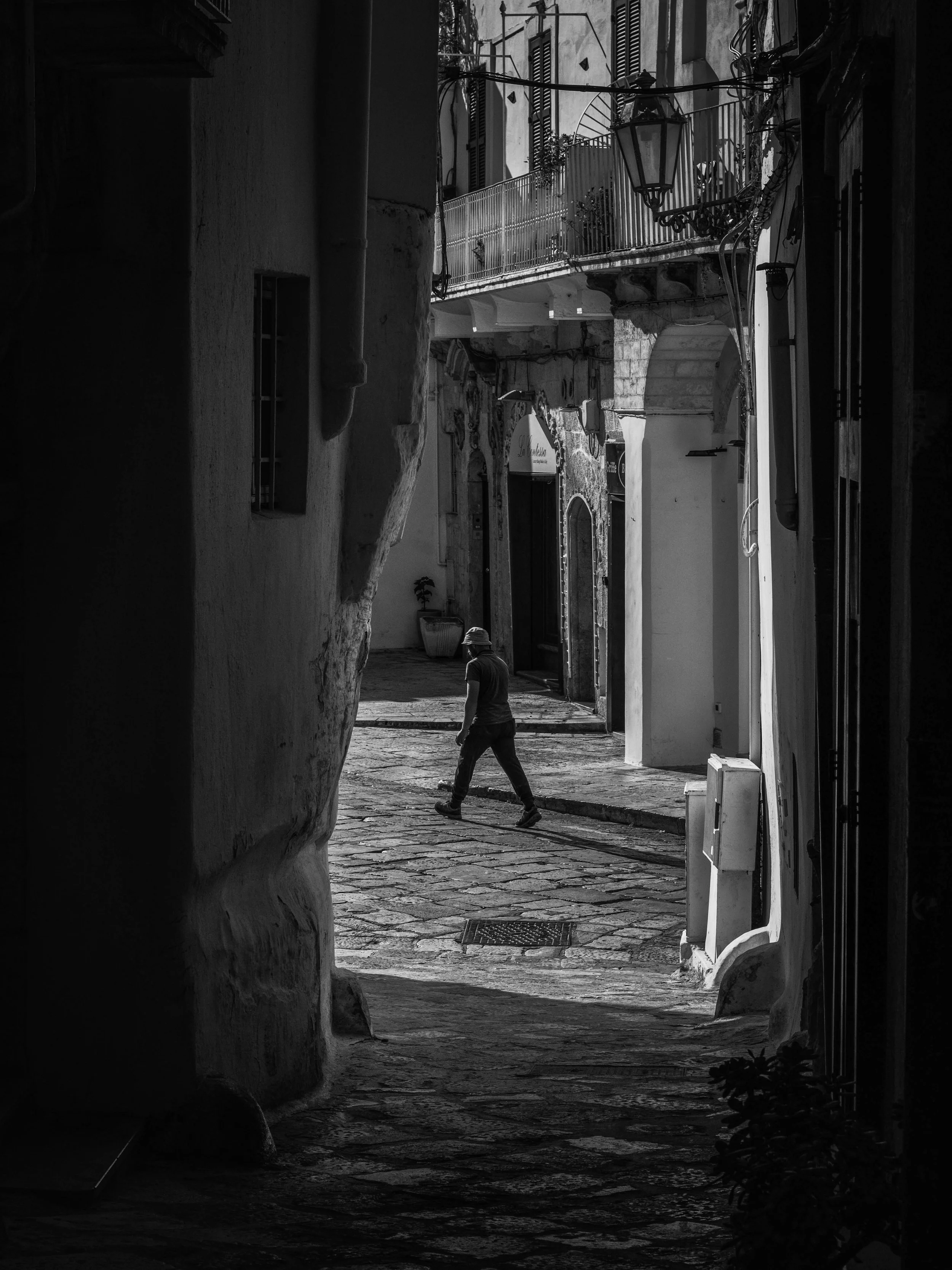 A narrow street scene in black and white, featuring a person walking away, old buildings with balconies, a street lamp, and cobblestone pavement.