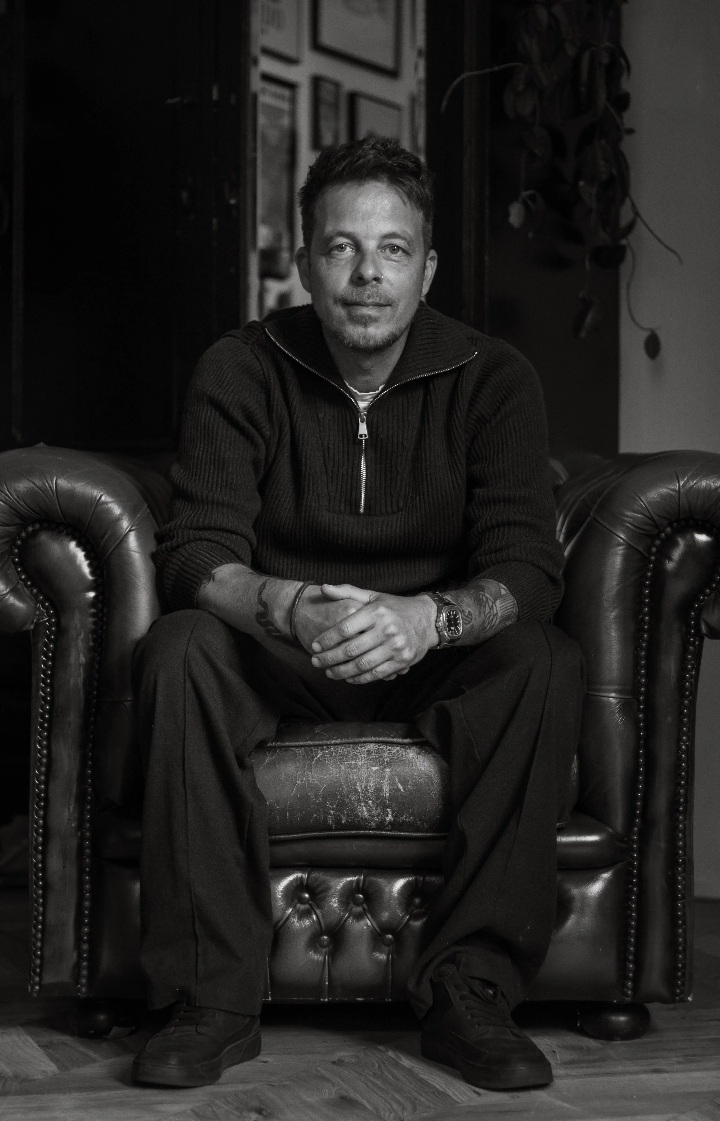 A man with tattoos and a beard sitting on a leather armchair in a room with framed photos on the wall.