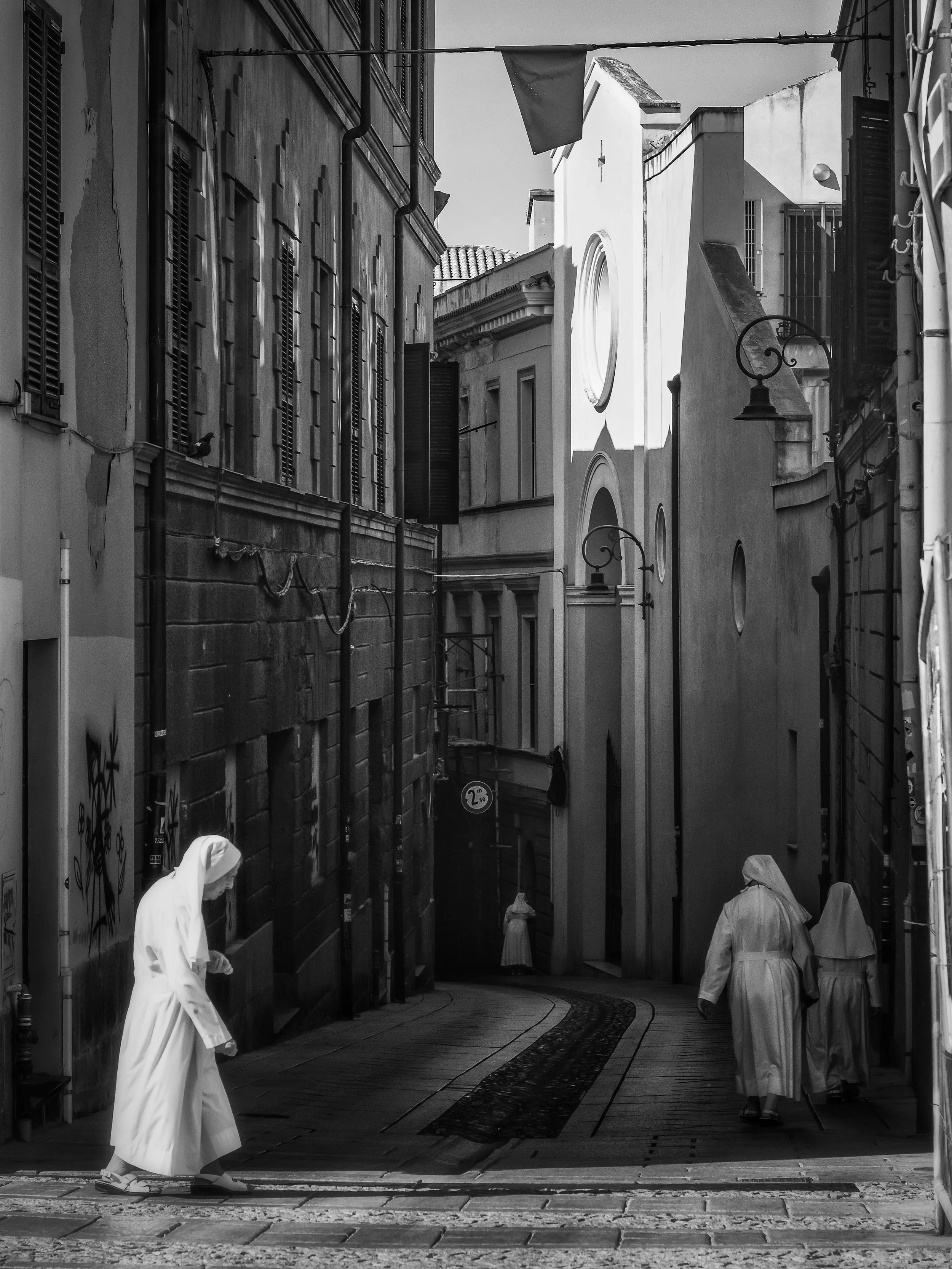Black and white photo of a narrow street with tall buildings on both sides. A person in a white robe and head covering is walking on the left, and three people in similar attire are walking away from the camera on the right. There are street lamps an