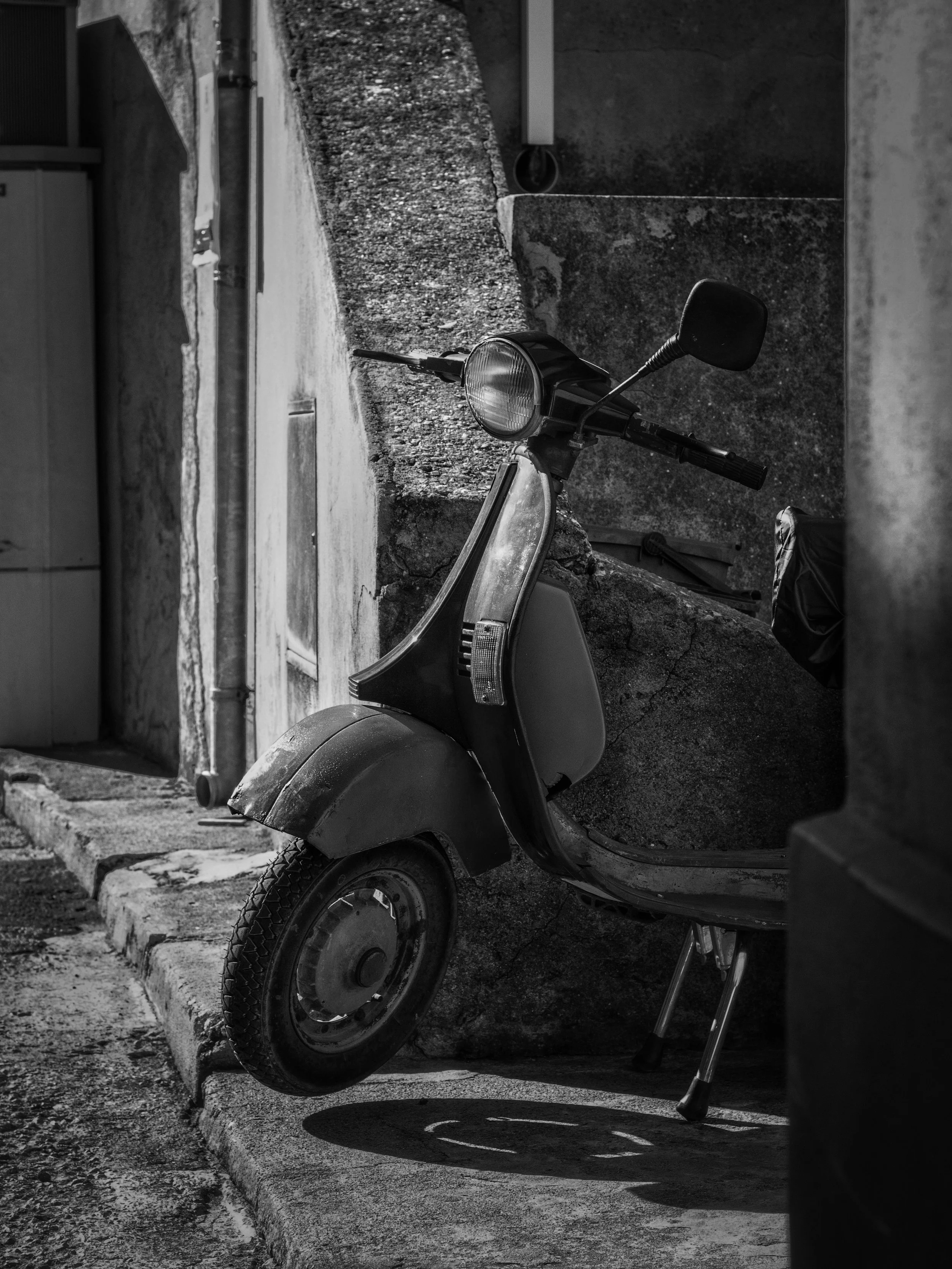 Black and white photo of a scooter parked outside, with a concrete curb and building in the background.