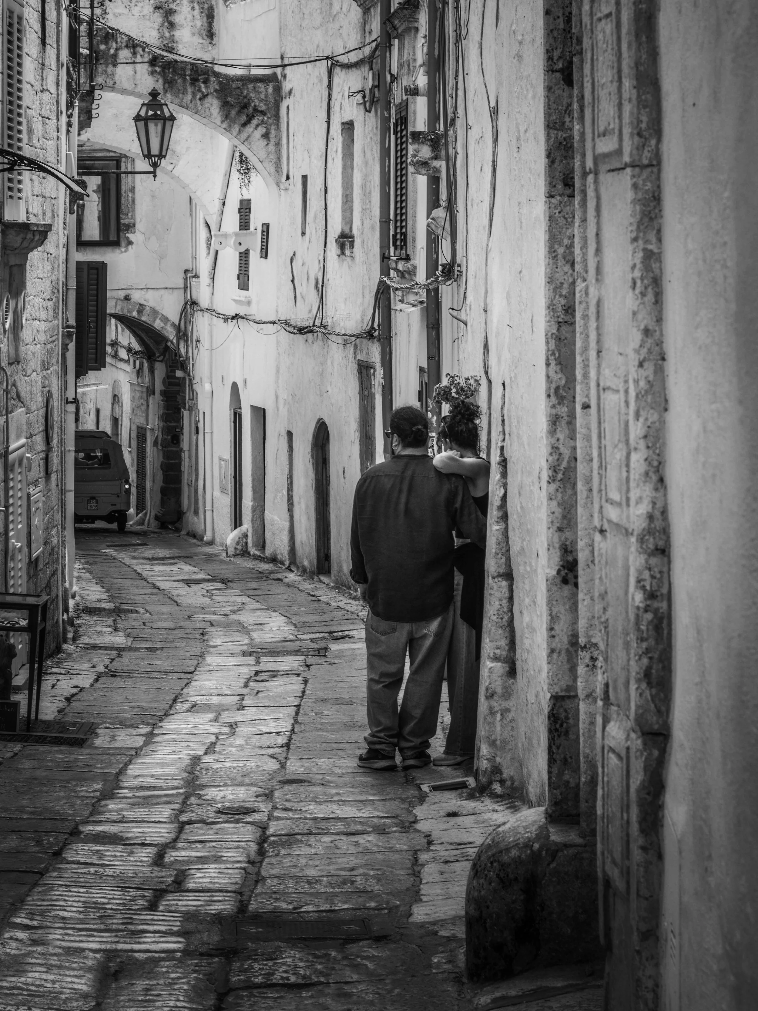 A narrow cobblestone street in an old town with stone buildings, a street lamp, and two people standing close together, one with glasses and the other with a bun, seen from behind.