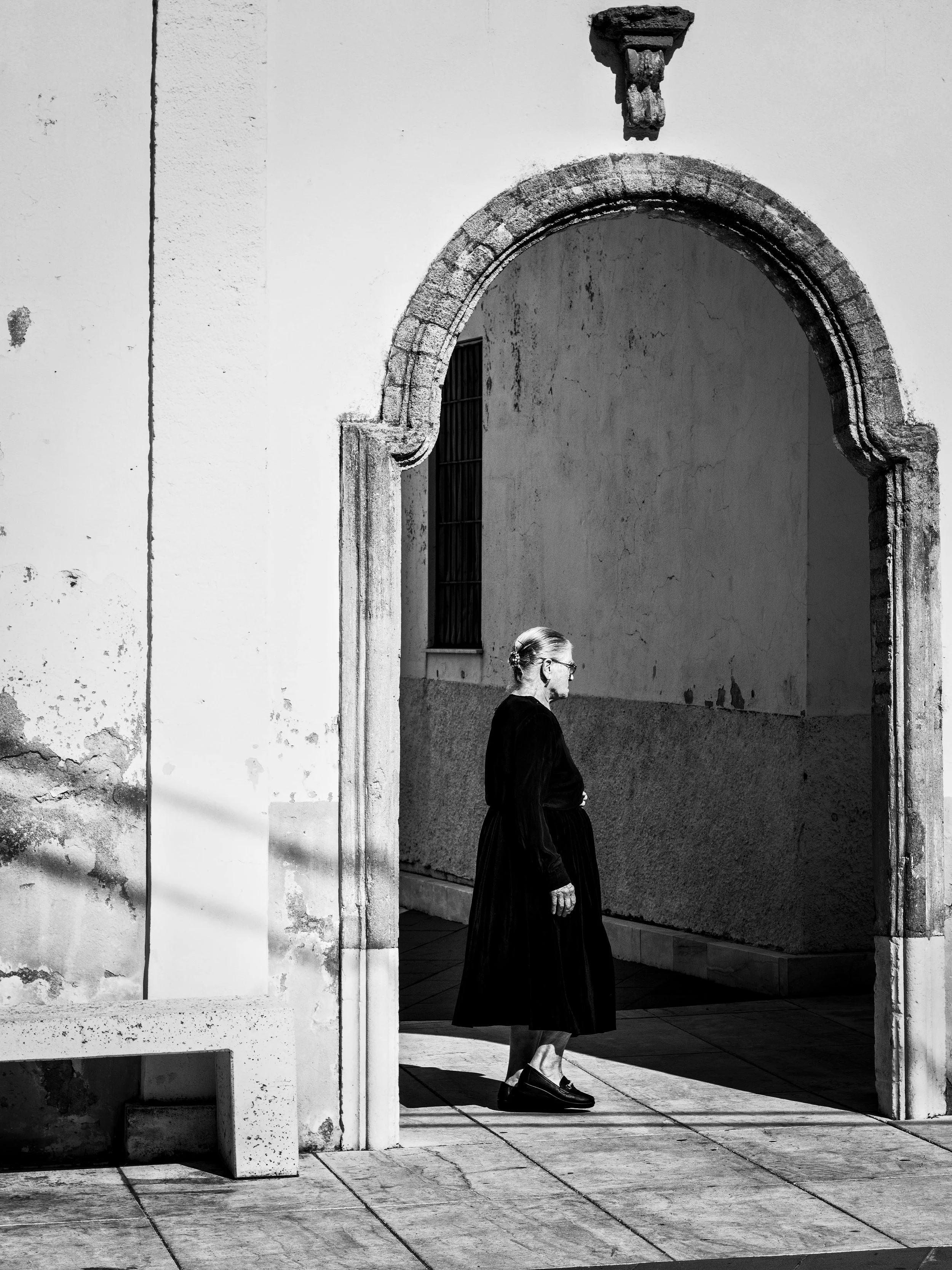 A black and white photo of a woman walking past a building with arched doorway and barred window. She is dressed in dark clothing and wears glasses, with her hair pulled back.