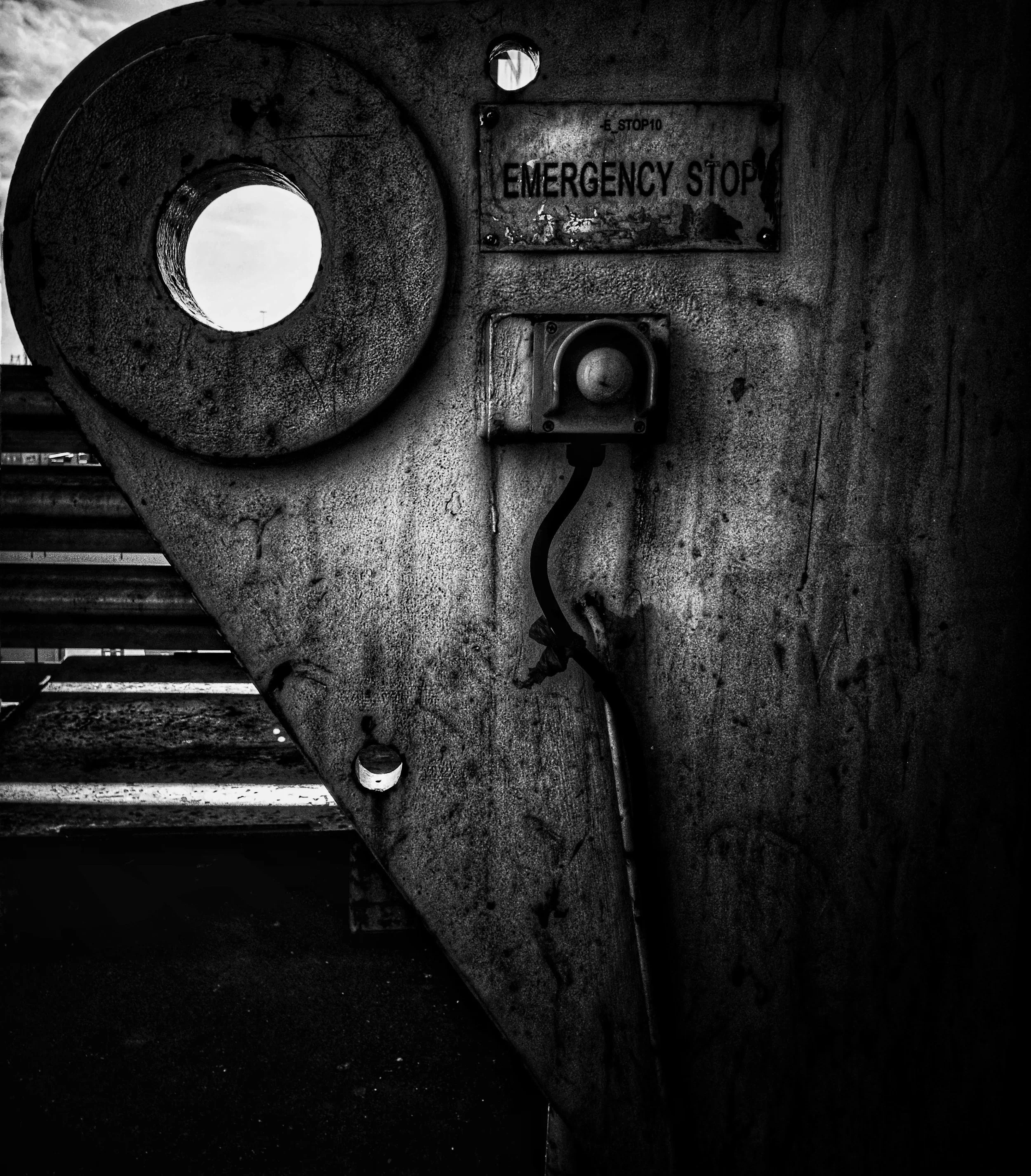 Black and white photo of an industrial or outdoor scene featuring a large circular pipe or structure with a hole in its side, a worn sign that reads 'Emergency Stop,' and an electrical box with a round protrusion on its cover, all mounted on a textur