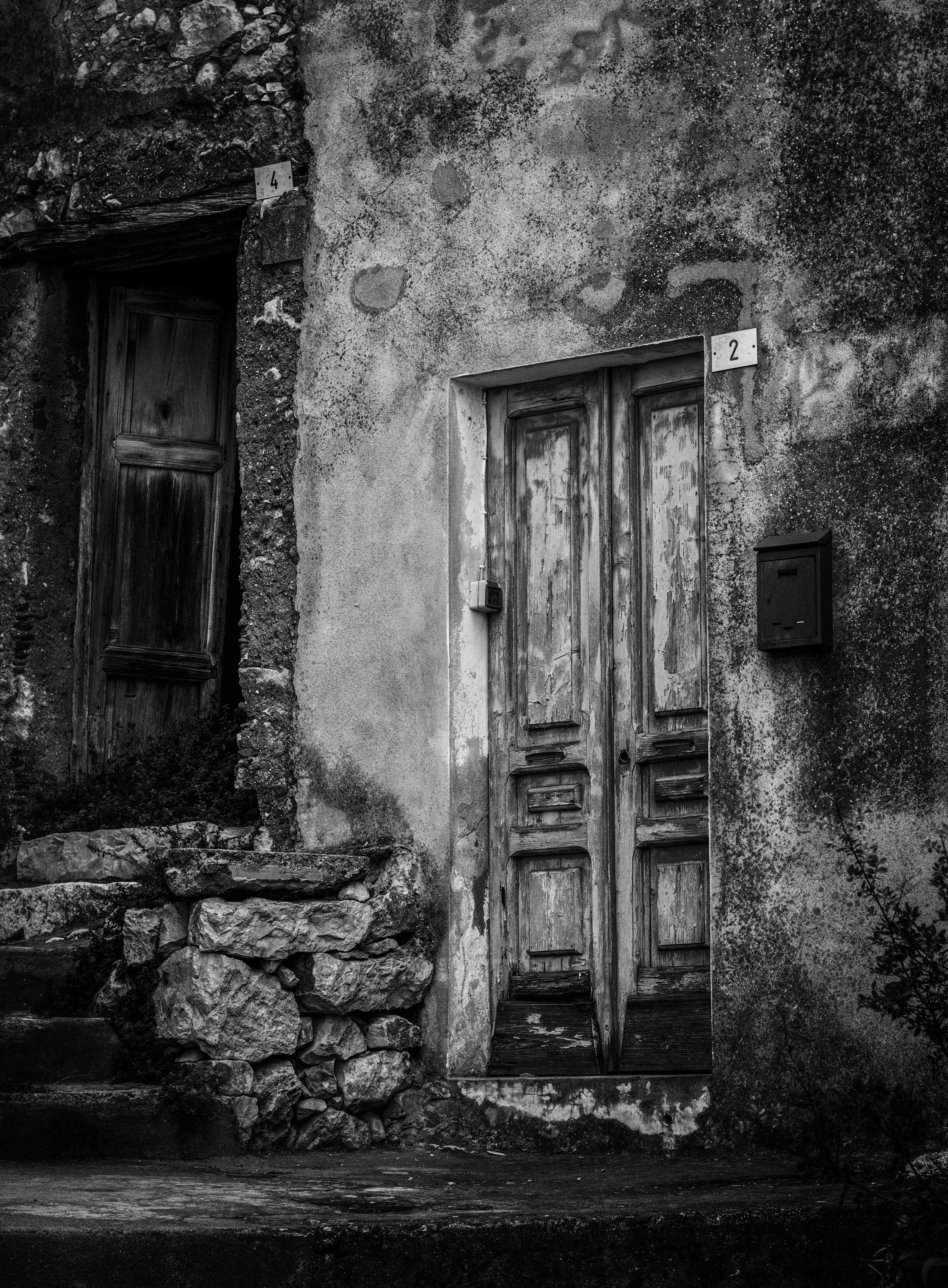 Black and white photo of an old, weathered door with peeling paint, set into a wall that has textured stucco and exposed stone on the left. Two small signs with the number 4 and 2 are mounted above the doors. A mailbox is attached to the right of the
