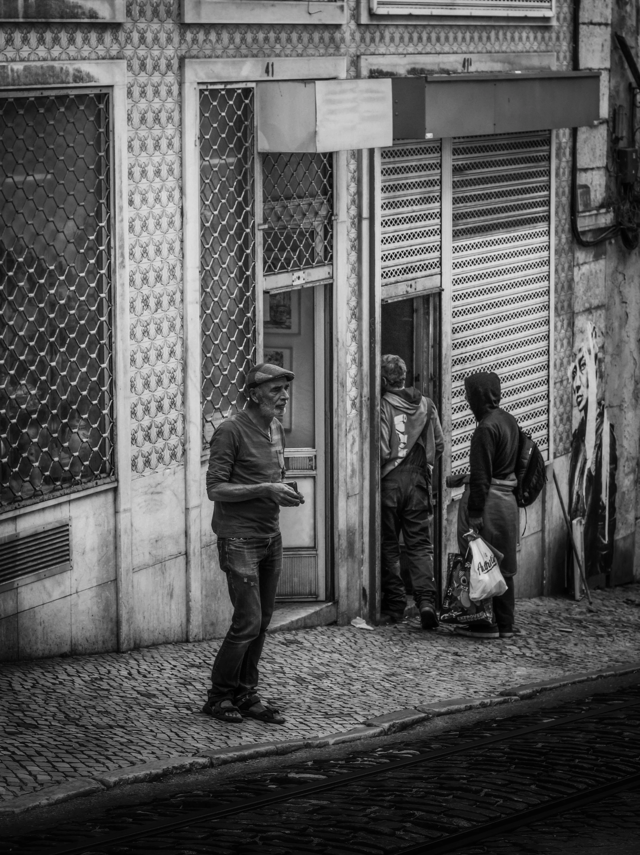 A black and white photo of a street scene with three people walking along cobblestone pavement in front of a building with barred windows and closed metal shutters. One man is standing and looking to his right, while two others walk towards a doorway