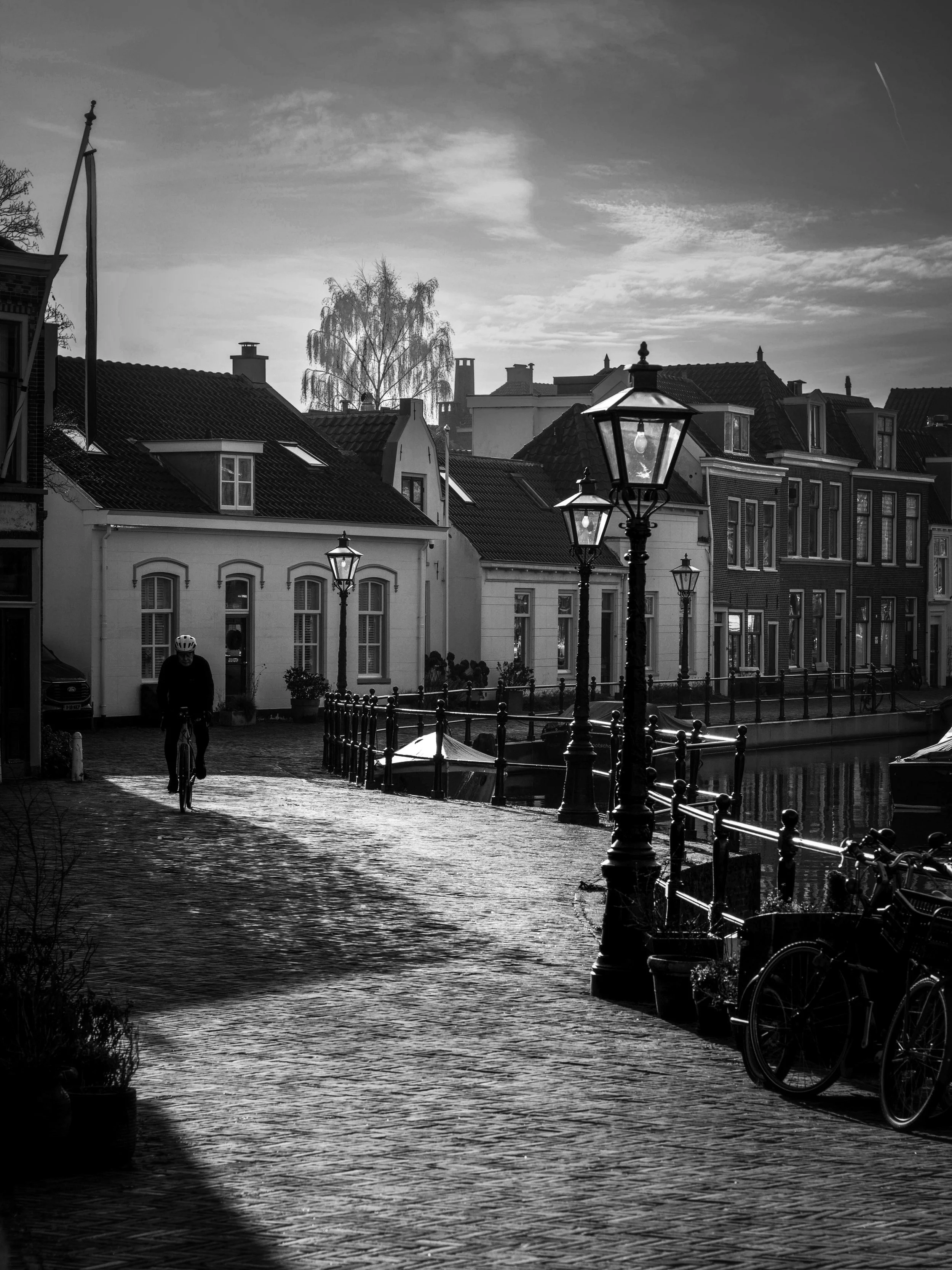 A black and white photo of a lakeside street with old-fashioned street lamps, waterfront houses with pitched roofs, a cyclist riding along the cobblestone path, and a person on a bicycle under a cloudy sky.