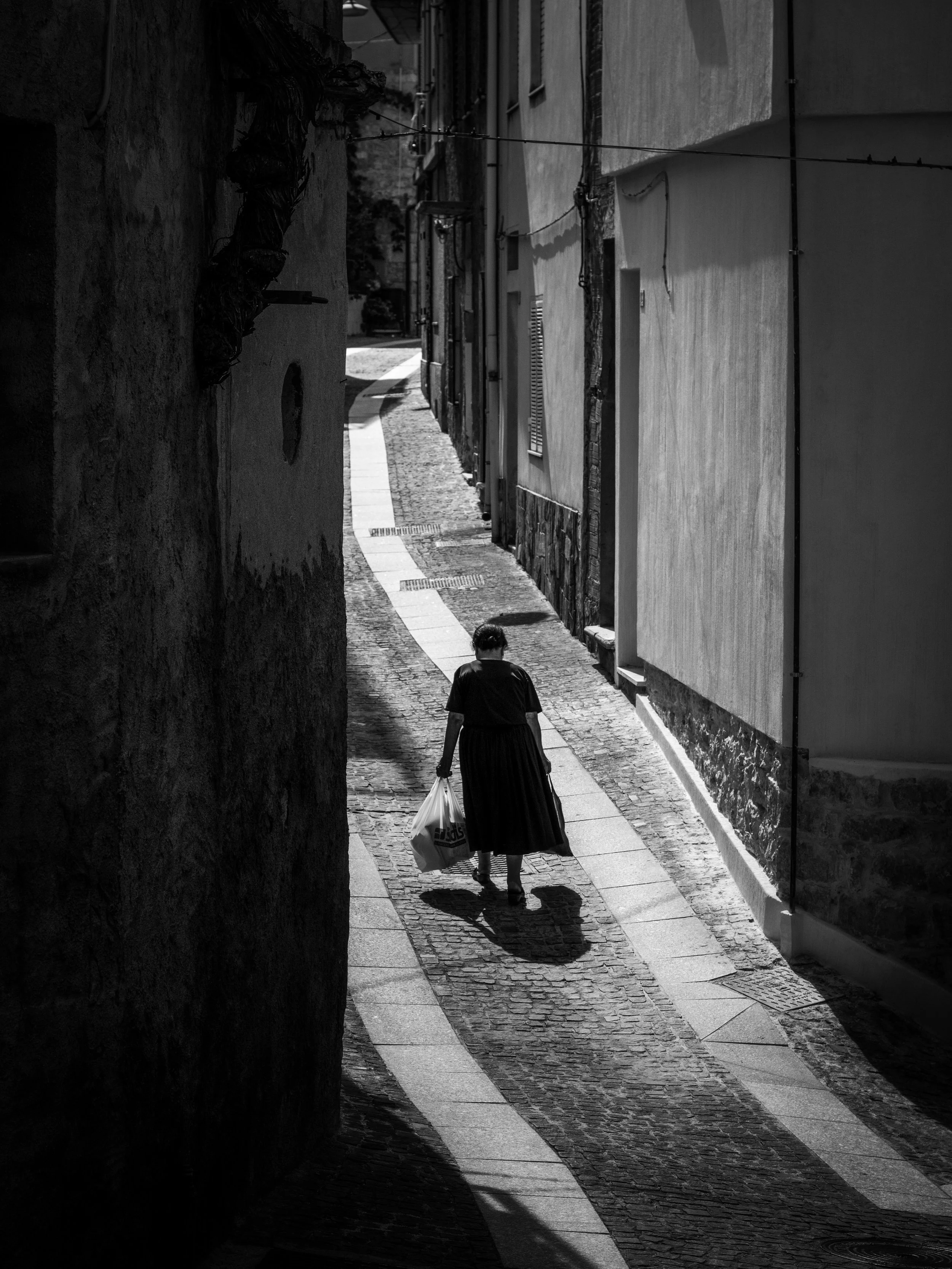 A woman walking down a narrow, cobblestone alleyway carrying shopping bags in both hands. The scene is illuminated by sunlight, casting her shadow on the ground. The alley is flanked by buildings with visible windows and ventilation panels.