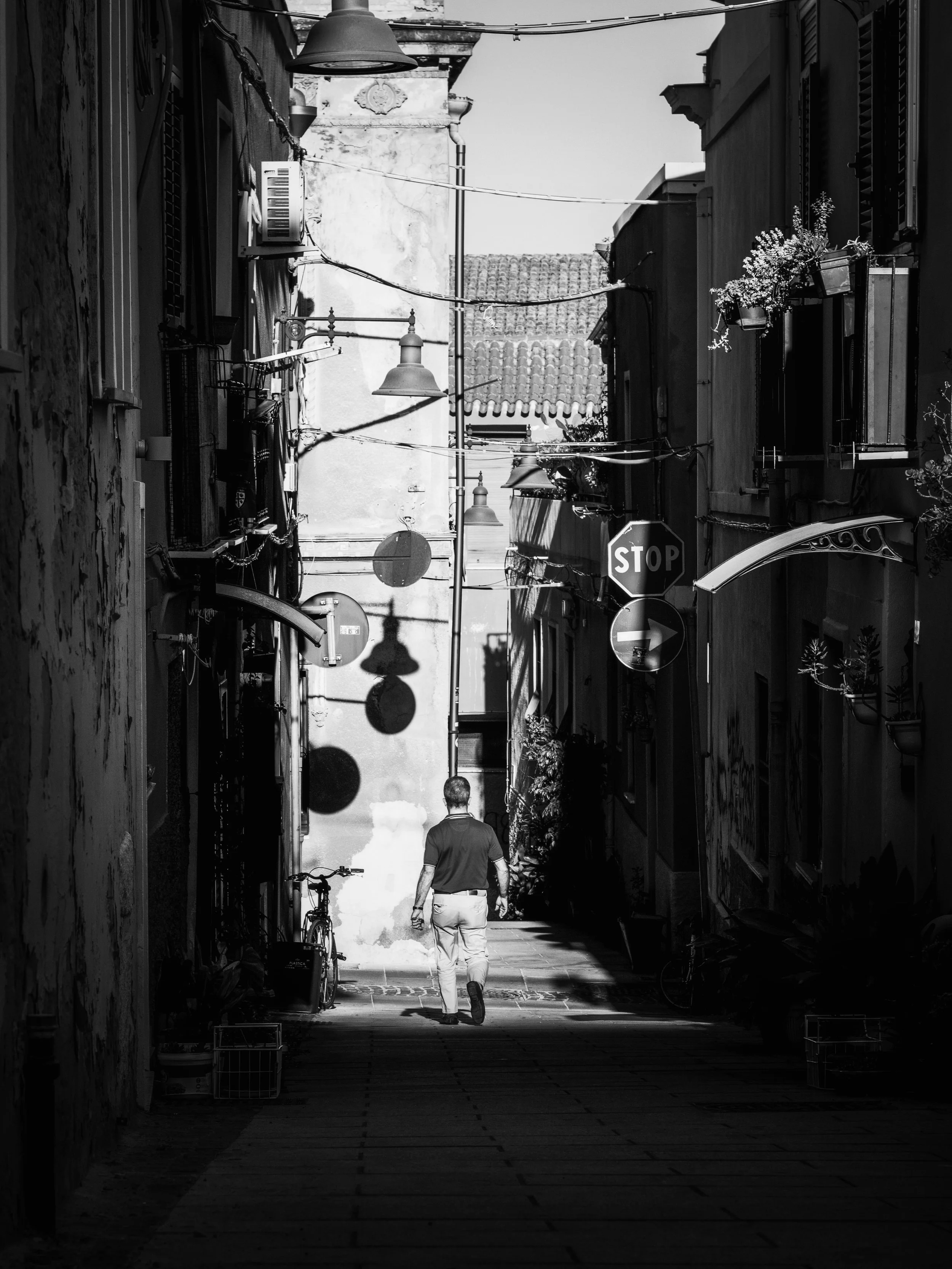 A black and white photo of a narrow alleyway with overhead street lamps, hanging flower pots, and a stop sign. A man walking away from the camera and a bicycle are visible.