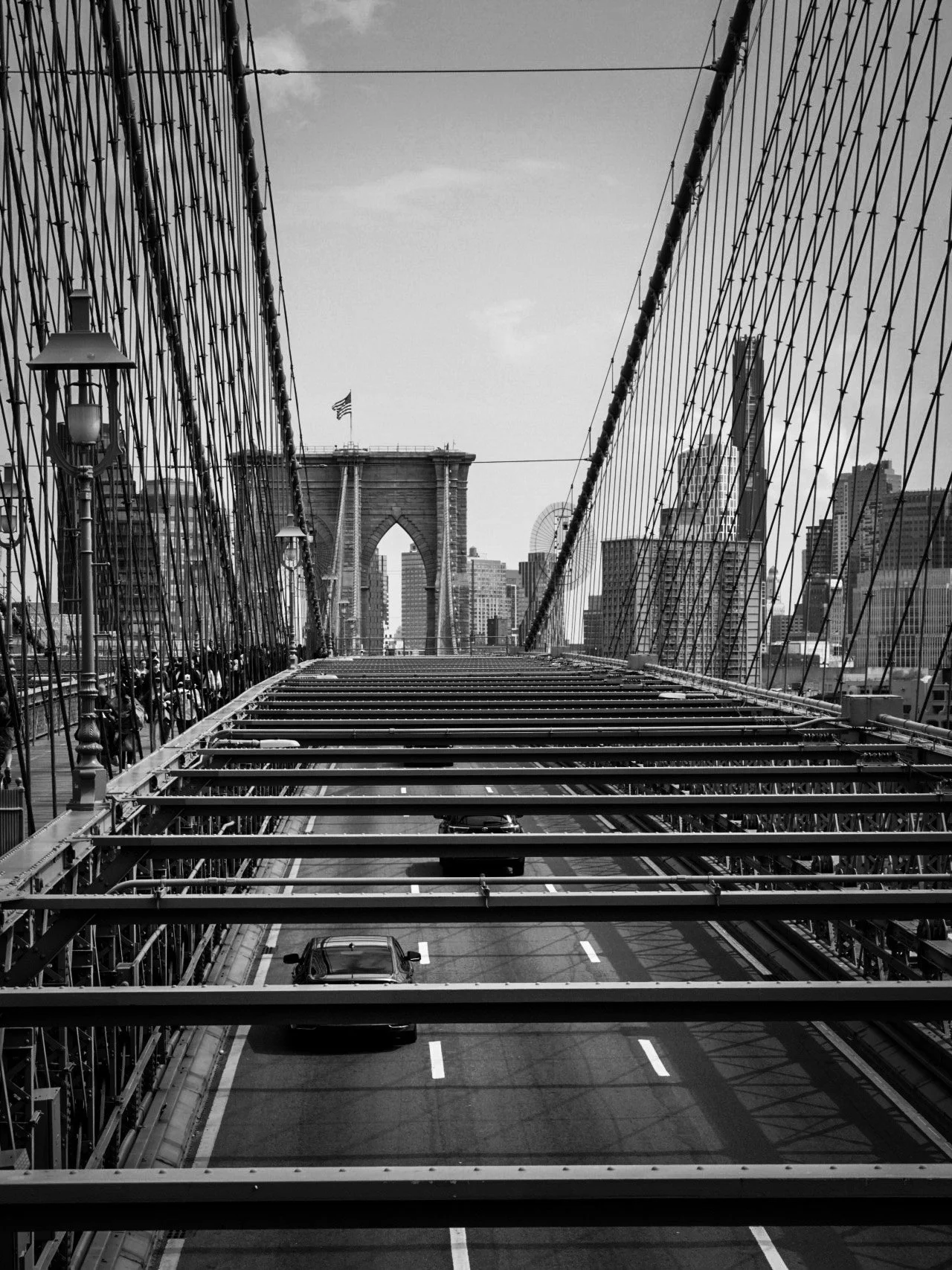 Black and white photo of the Brooklyn Bridge showing pedestrians walking on the bridge and cars driving underneath on the road, with the Manhattan skyline in the background.