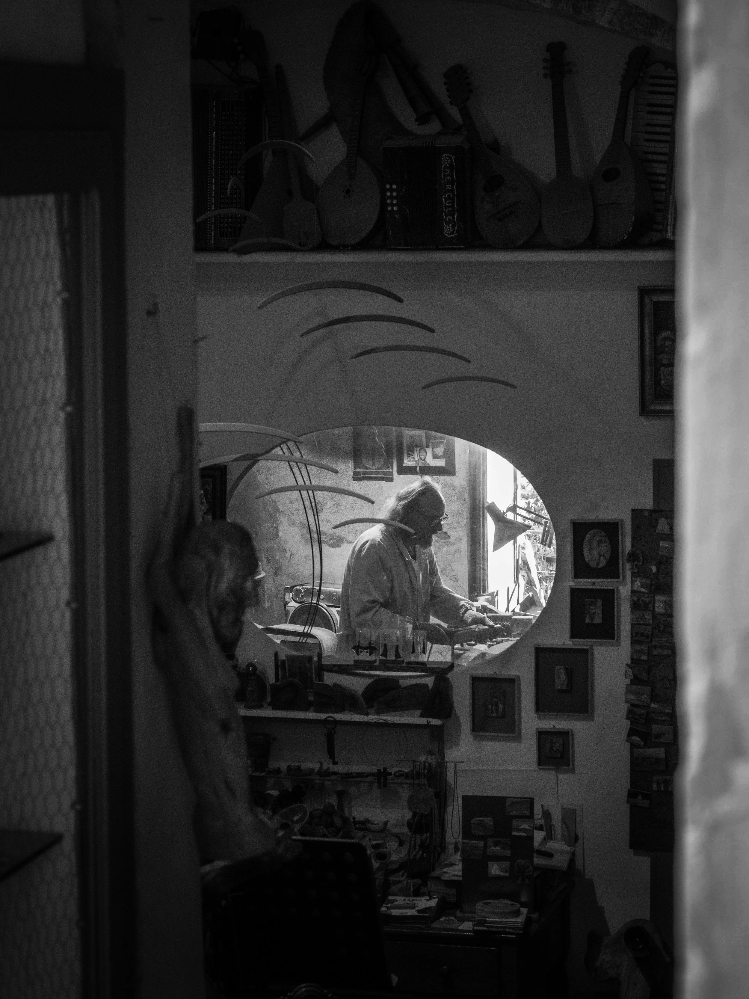 A black and white photo of a man playing a keyboard or piano in a room, with various guitars hanging on the wall in the background. A woman and another person are partially visible in the foreground, and there are pictures and objects on the walls an