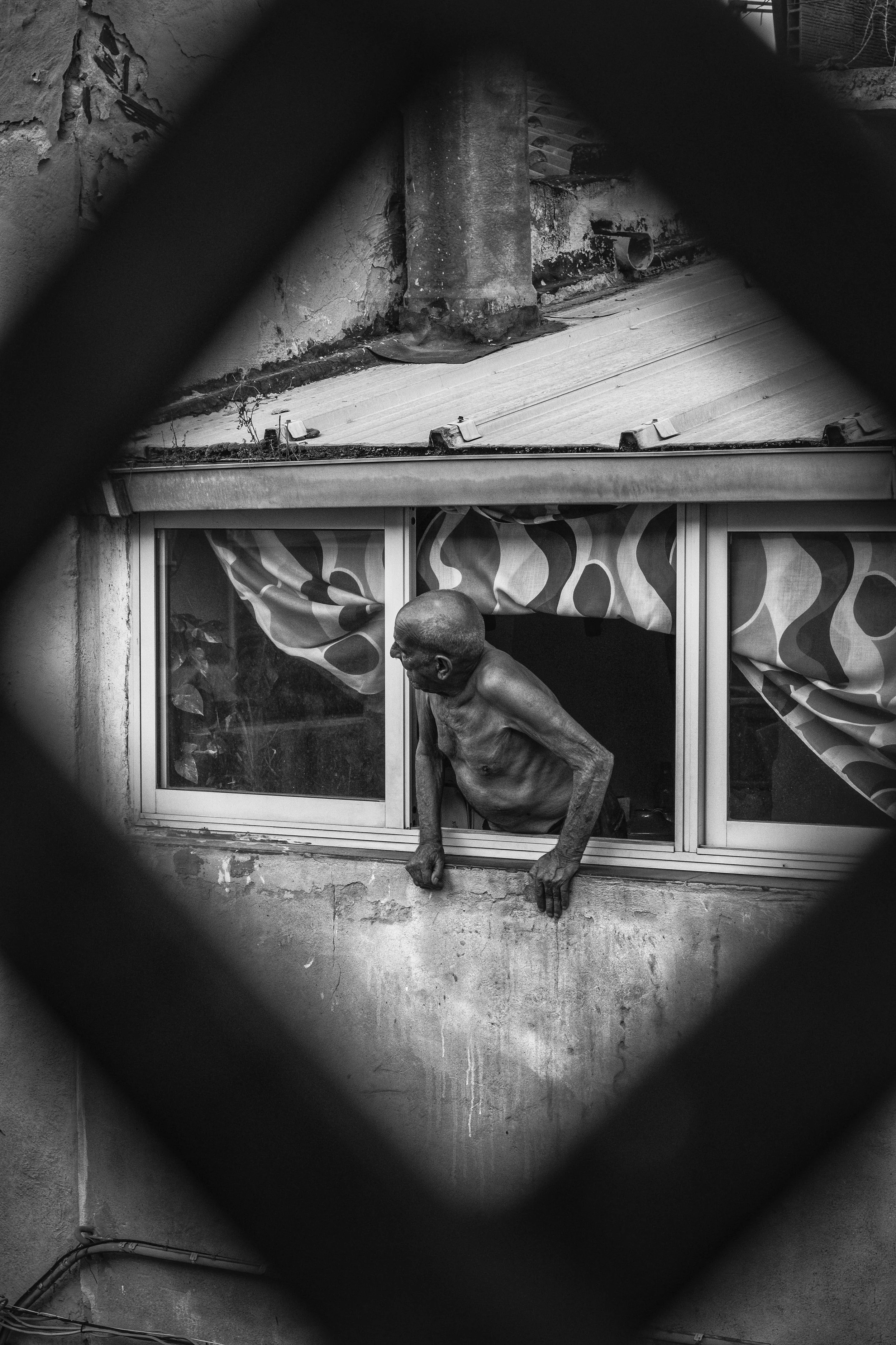 A shirtless elderly man leaning out of a window, viewed through decorative window frames, with a weathered building background.