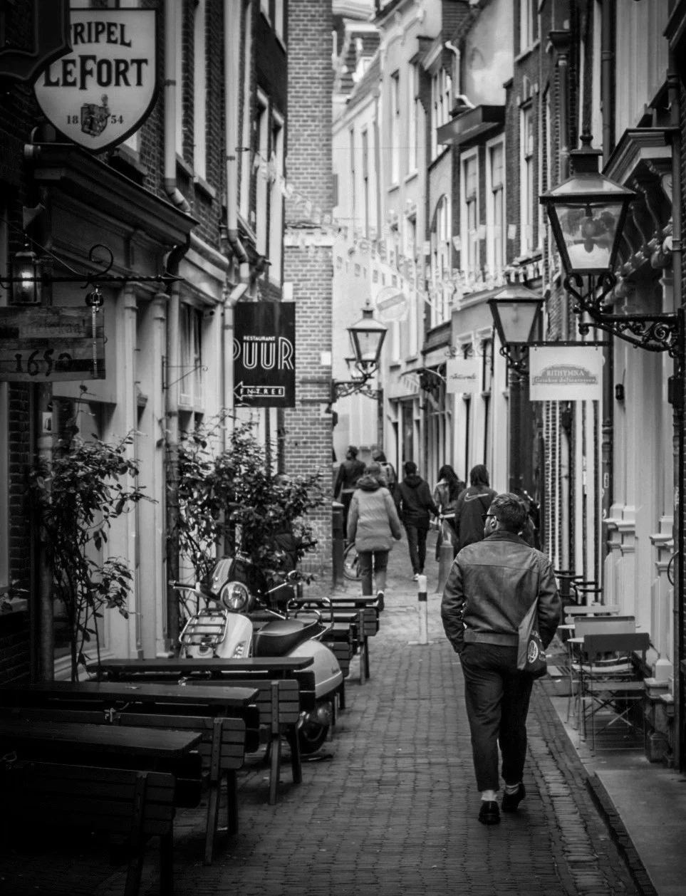 A black and white photo of a narrow European street with cafes, shops, benches, and street lamps. Pedestrians walk along the cobblestone path.