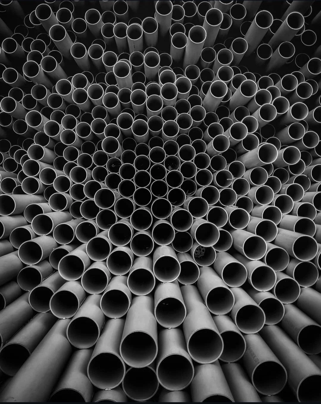 A black and white photo of numerous metal pipes arranged in a circular pattern, viewed from the front showing their open ends.