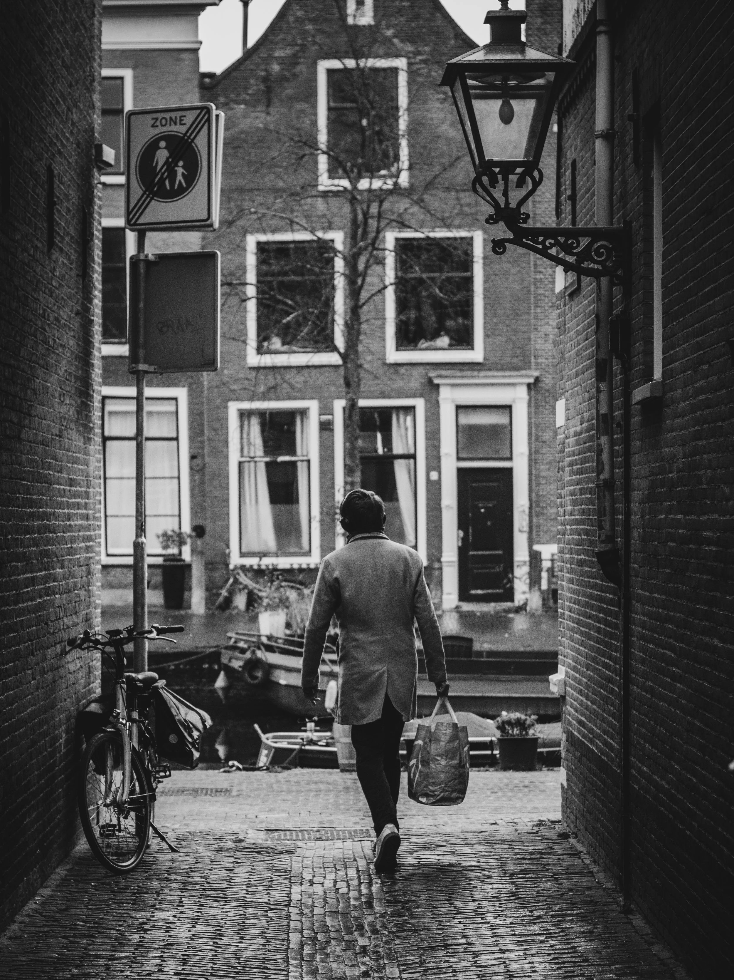 A man walks with a shopping bag in an alley between brick buildings; a bicycle leans against the left wall, and there are signs and a lamp on the right wall. The background shows a tree and residential buildings.