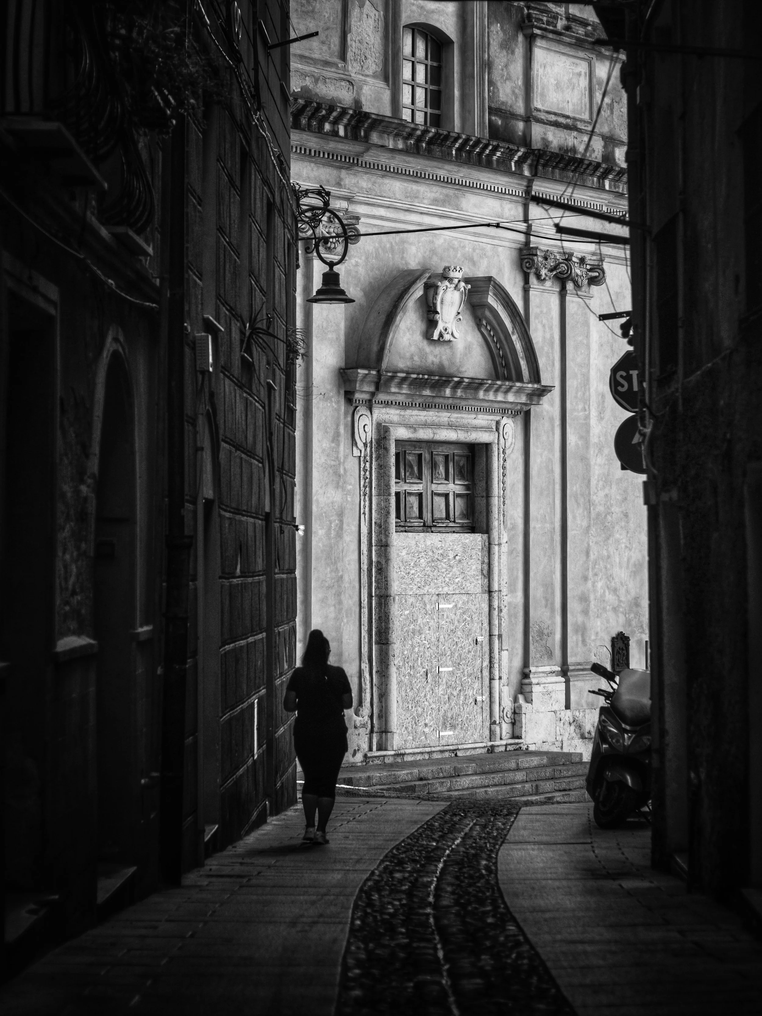 Black and white photo of a narrow street with a woman walking away, an old building with ornate architectural details, a motorcycle parked on the right, and a stop sign on the right side.