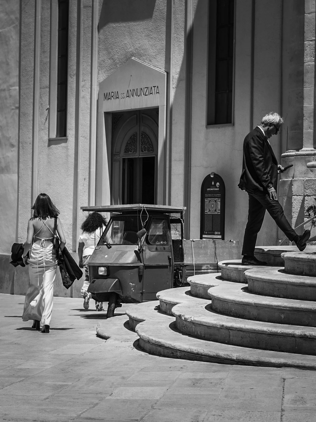 People walking and a man on stairs outside a church with a sign reading 'Maria SS Annunziata' in black-and-white
