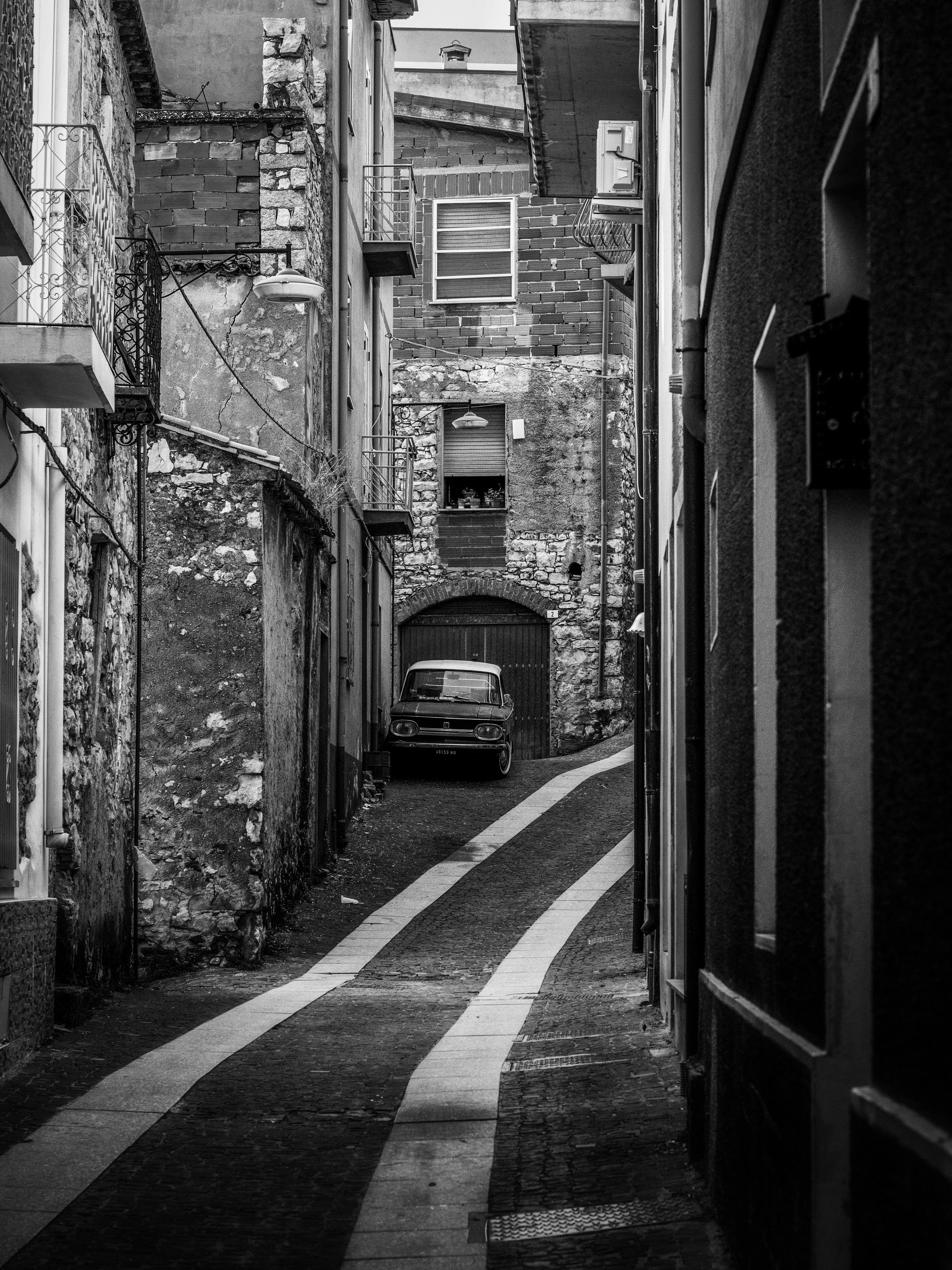 Narrow alleyway with textured, aged buildings on either side, a vintage car parked near a large door, and several windows and outdoor lights, in black and white.