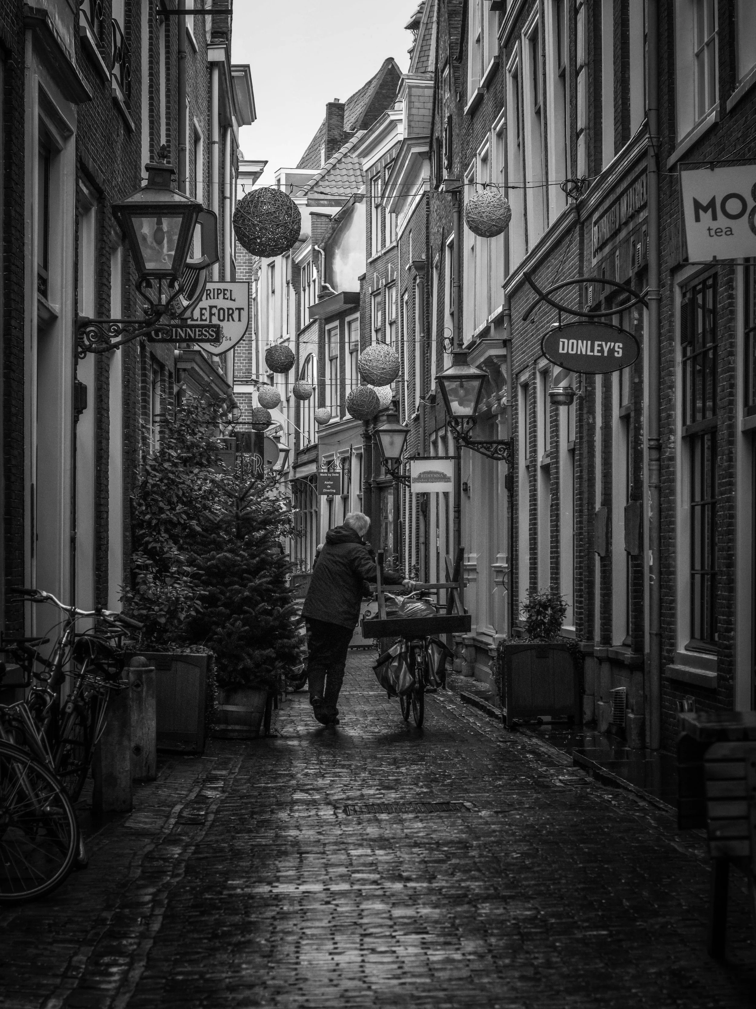A narrow cobblestone alleyway lined with brick buildings, decorated with hanging spherical lights, with a person walking a bicycle, and signs for pubs and coffee shops.