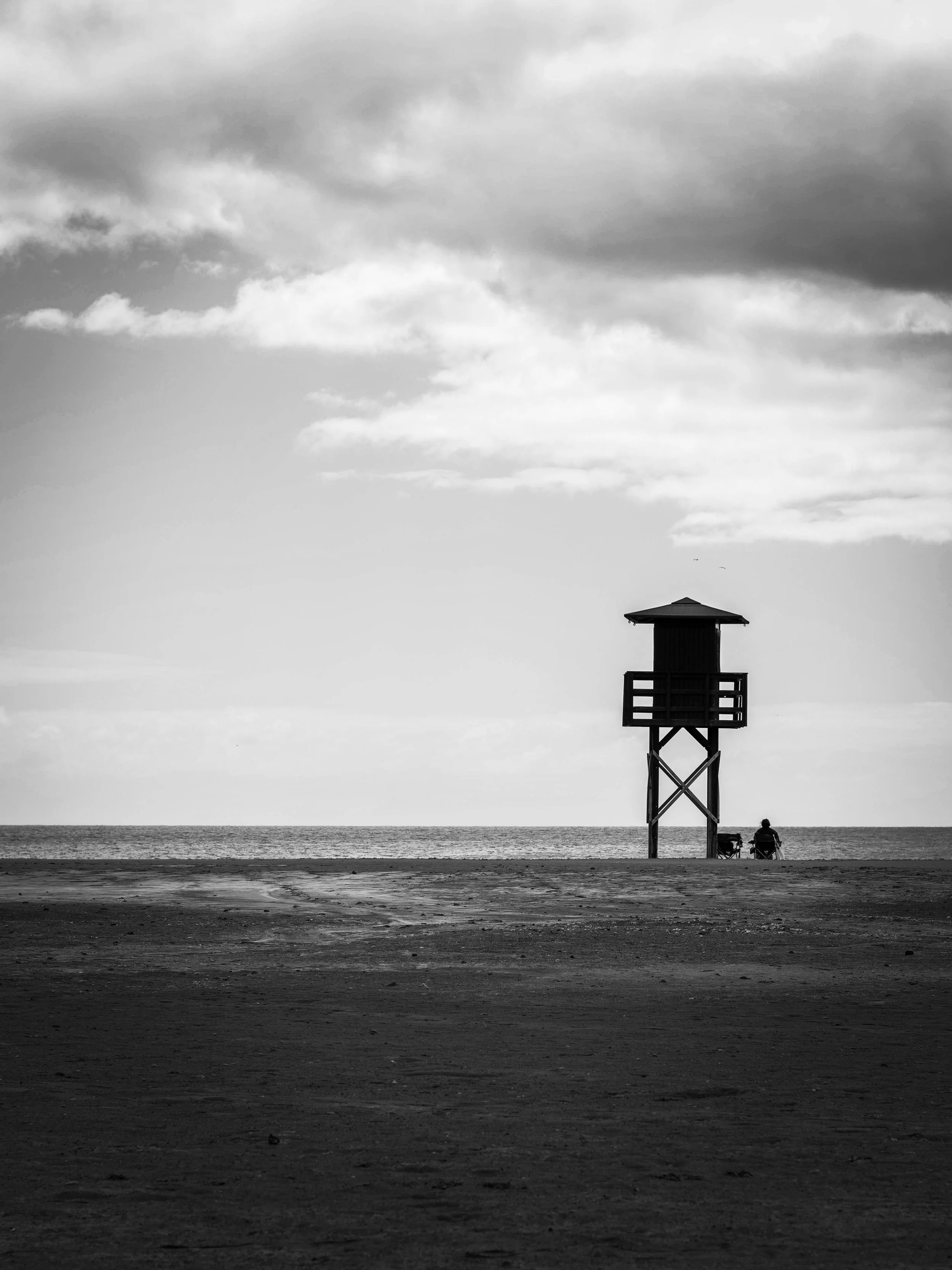 A black and white photo of a beach with two chairs and a lifeguard tower near the water, under a cloudy sky.