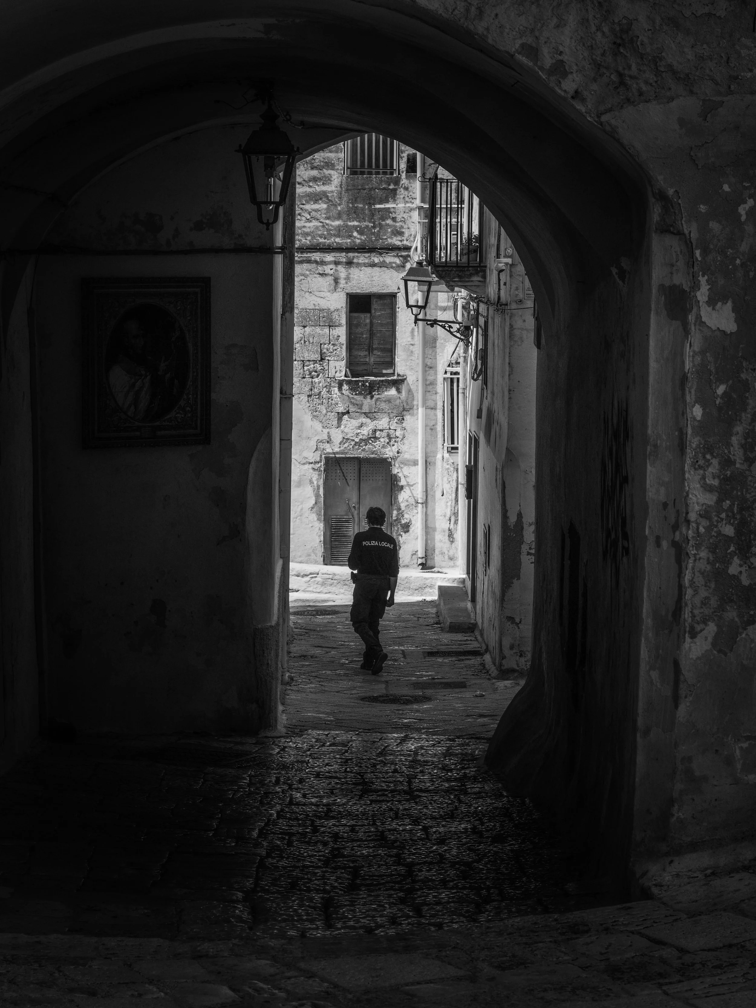 A man in a uniform labeled 'Polizia Locale' walking through a dark, arched alleyway in an old European city, with weathered buildings and hanging lanterns.