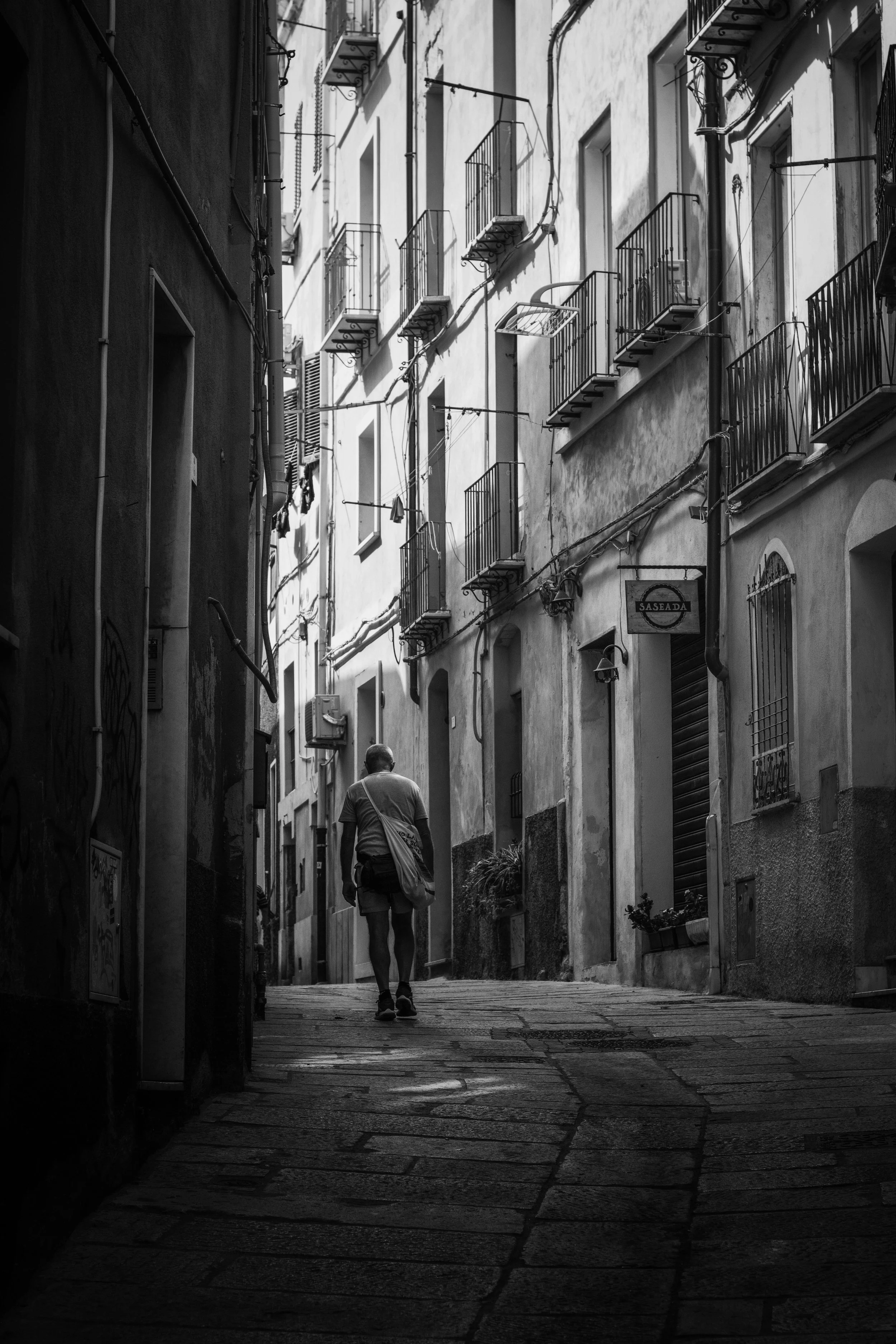 Black and white photo of a narrow alleyway with old buildings, small balconies, and a person walking away from the camera.