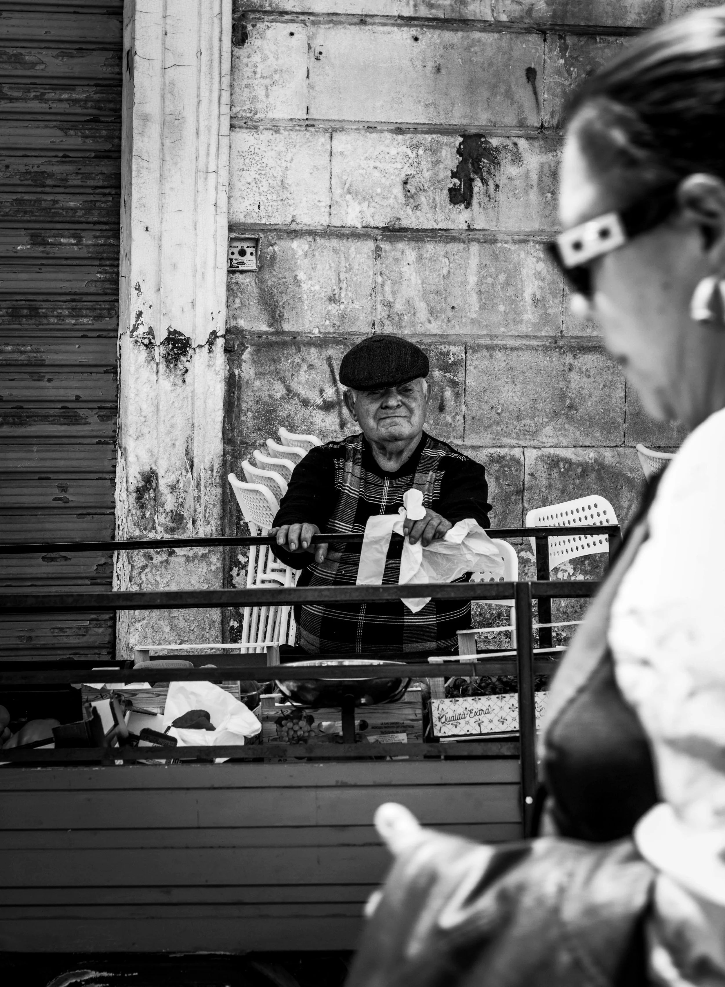 A black and white photo of an older man with a cap sitting behind a table with various items, looking at a woman in sunglasses and a mask standing in front of him. The background features a textured wall with a power outlet.