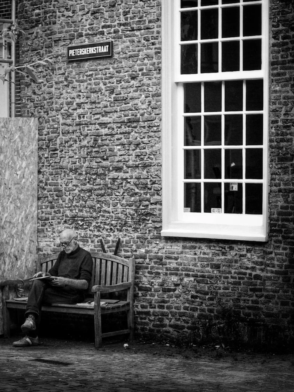 Man reading a book on a bench outside a brick building with a large window and a street sign that reads "Pieterskerkstraat."